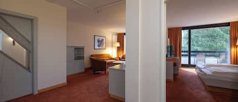 Quarto de hotel com cama, escrivaninha, cadeira, sofá e uma grande janela com cortinas.