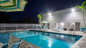 Una piscina con sillas de descanso y sombrillas, rodeada por una valla blanca y un edificio con puerta y ventanas.