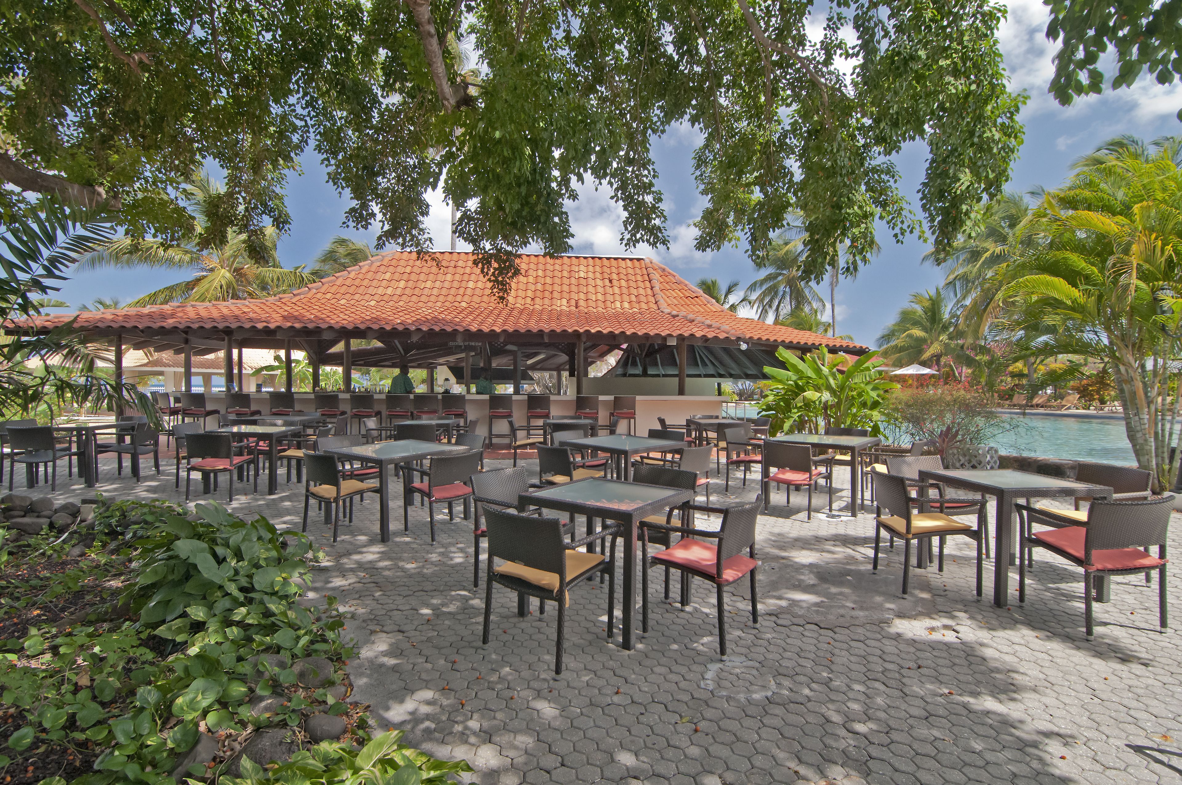 An outdoor patio area with tables and chairs, a covered seating area, and a pool in the background.