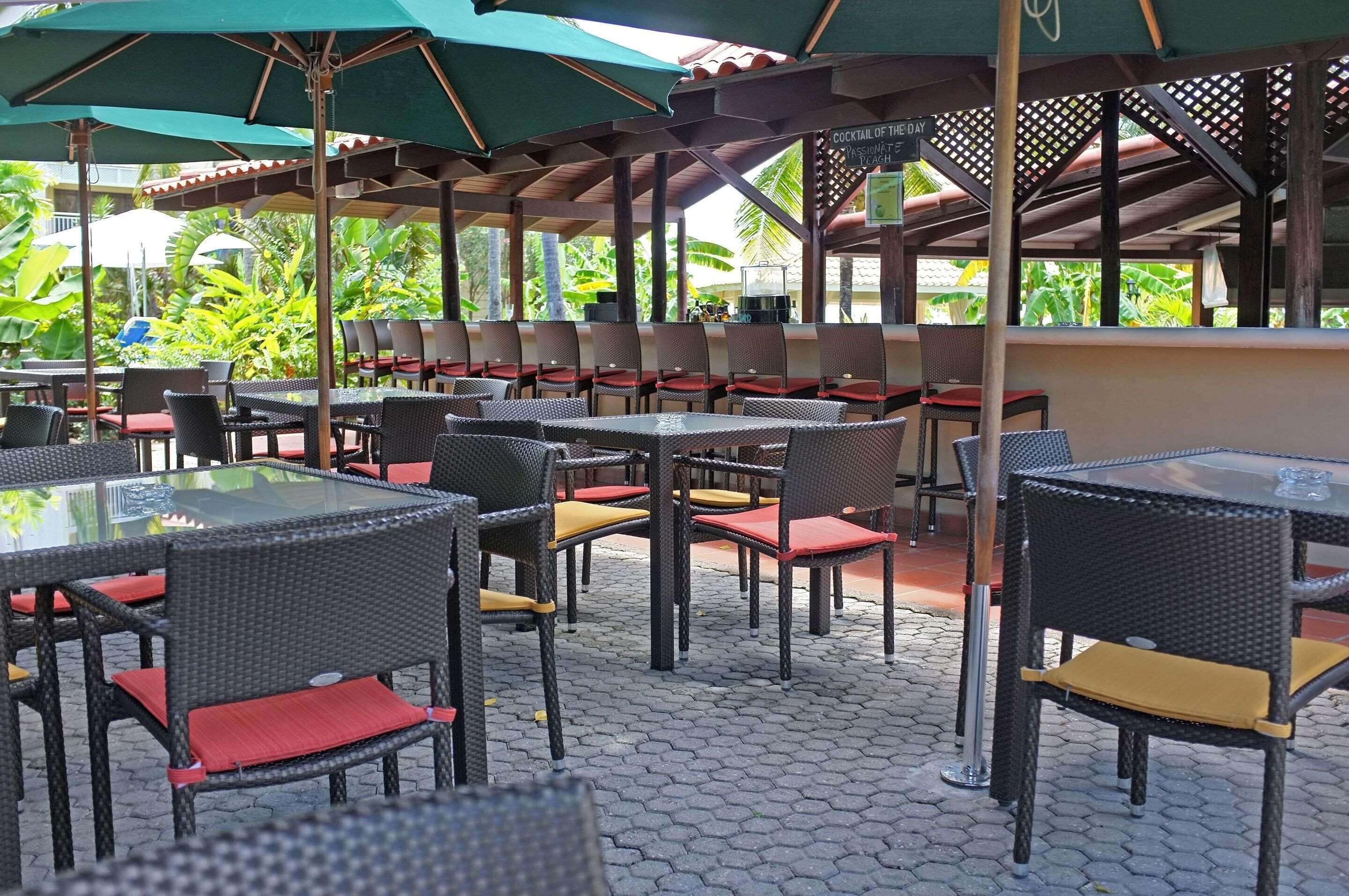 Un'area patio esterna con tavoli e sedie in vimini, alcune con cuscini rossi, sotto una struttura coperta con un cartello che indica 'Cocktail del giorno'.