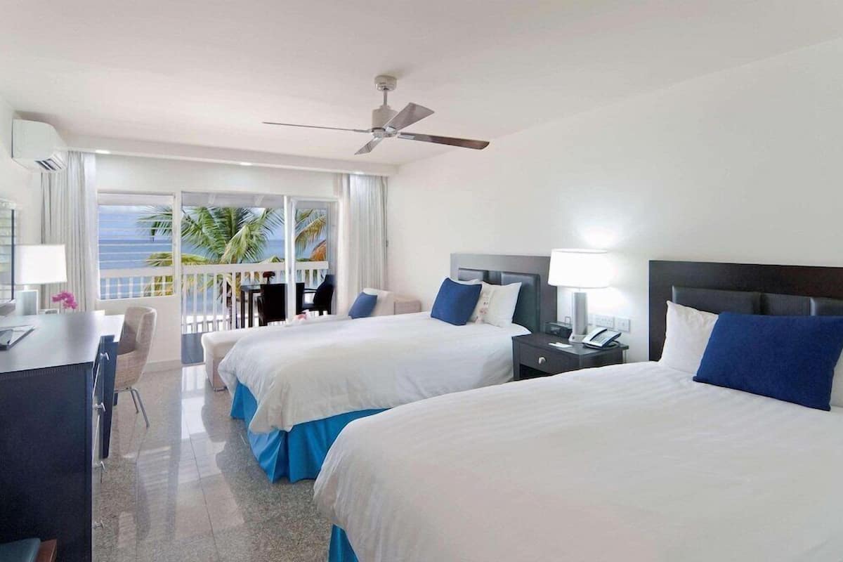A hotel room with two beds, a desk, a chair, a balcony with a view of palm trees, and a ceiling fan.