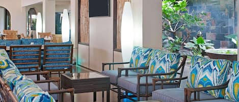 A modern lounge area with wicker furniture, a glass coffee table, and a view of a garden with a stone wall and plants.