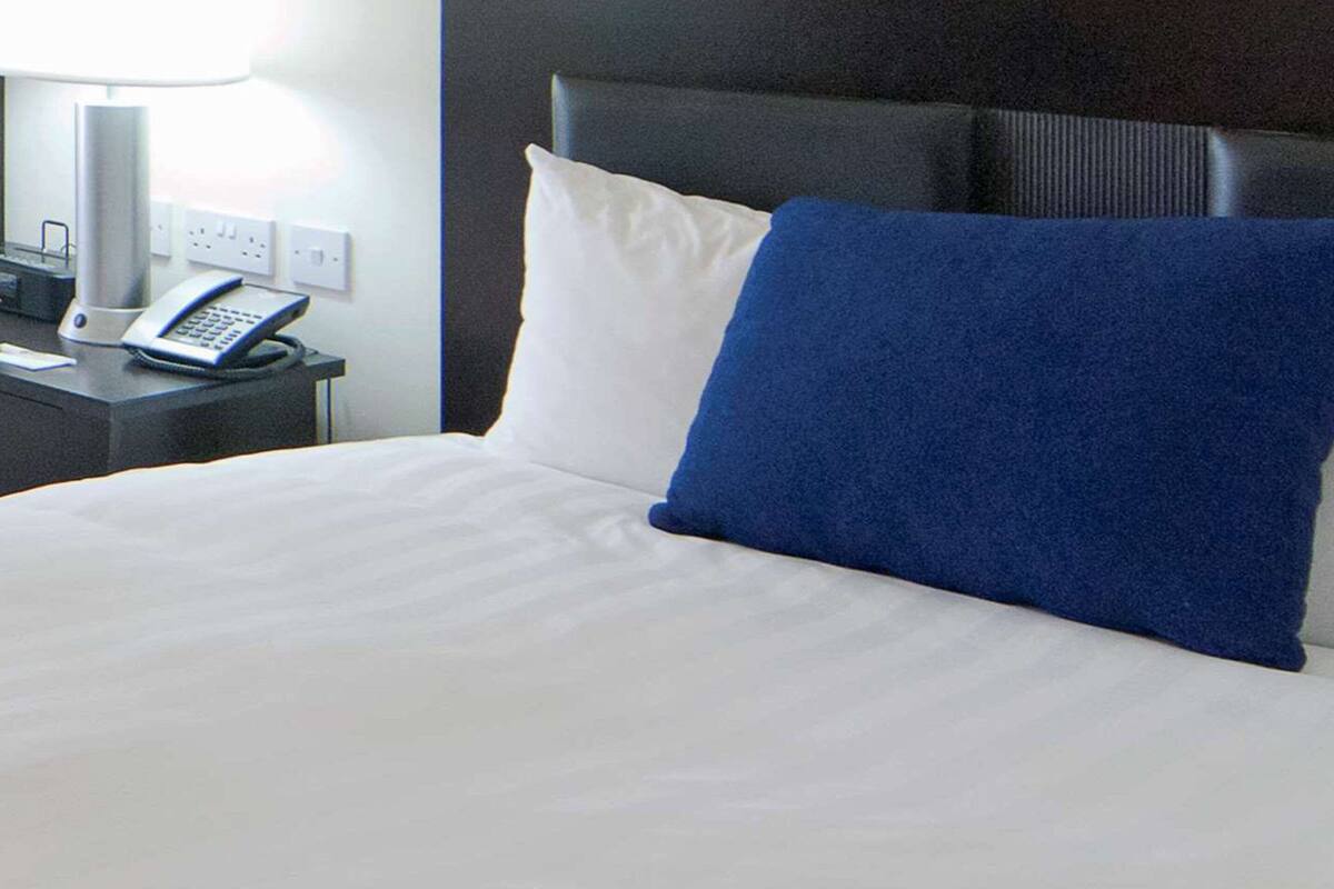 A neatly made bed with white and blue pillows, a bedside table with a lamp, and a telephone.