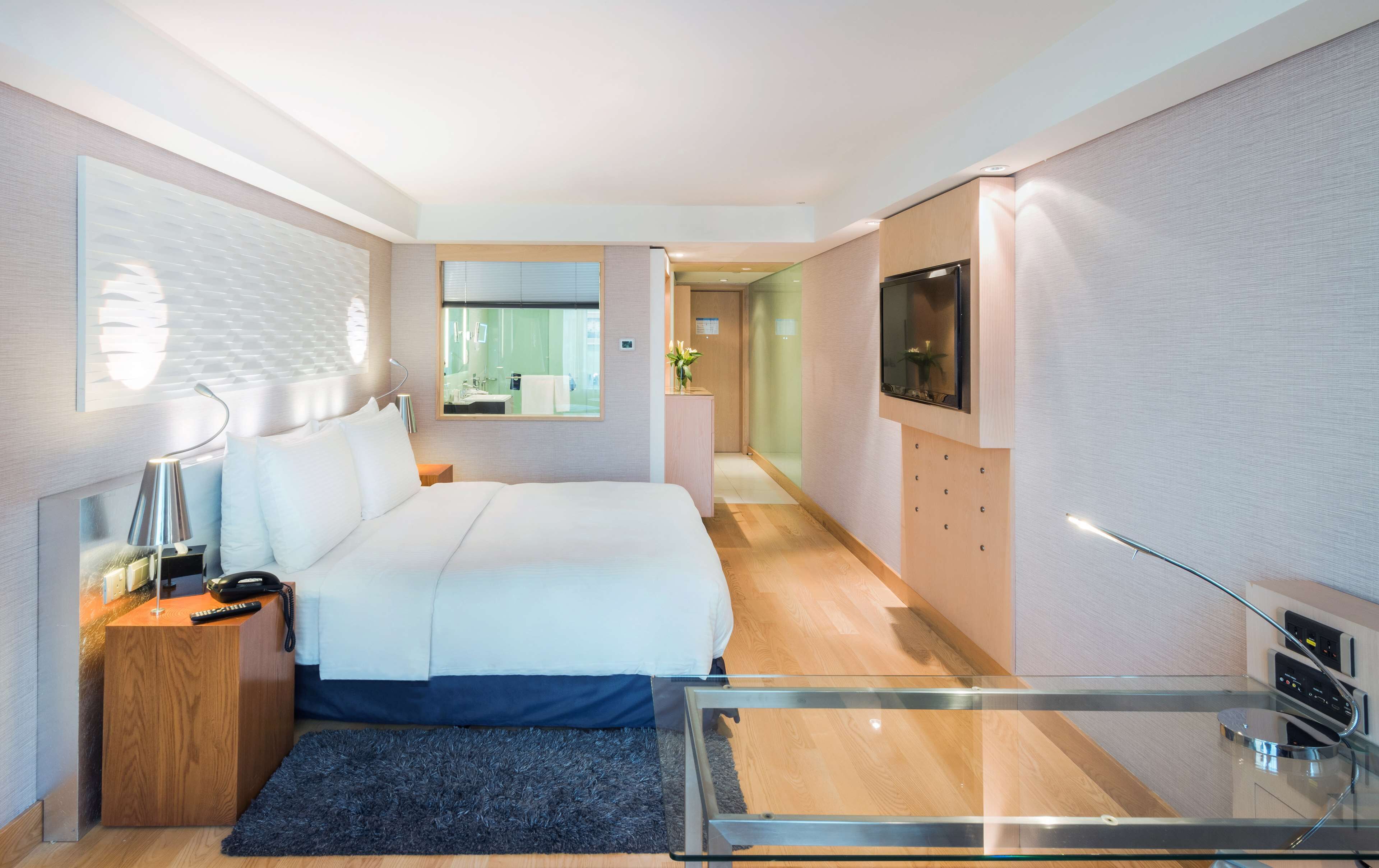 A modern hotel room with a large bed, a desk with a lamp, a TV, and a glass shower enclosure.