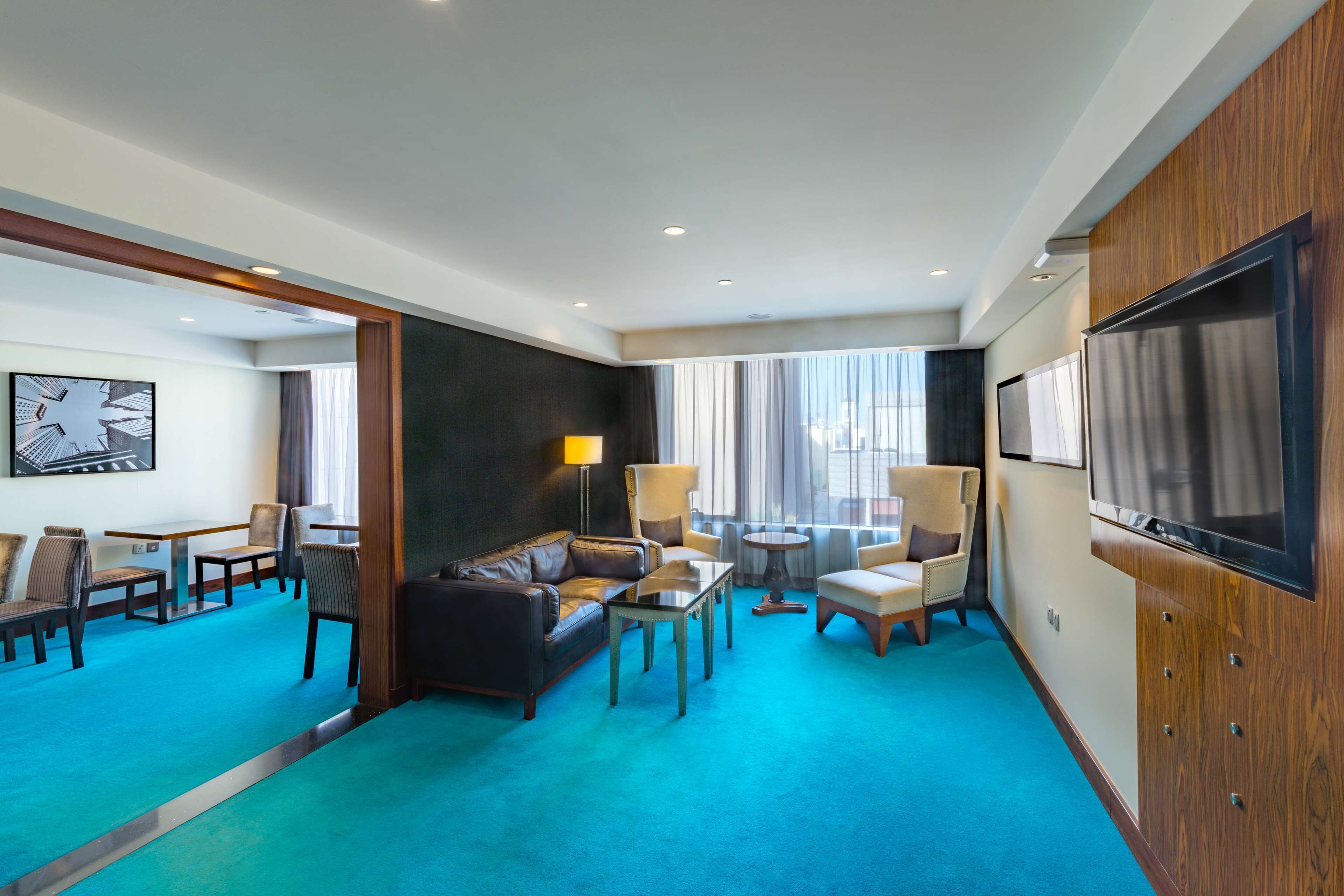 A hotel room with a living area featuring a sofa, chairs, a coffee table, a TV, and a large window with curtains.
