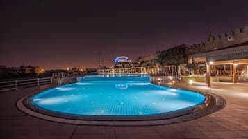 A swimming pool with a tiled deck, a modern building with a curved roof in the background, and a stone wall with a flag on top.