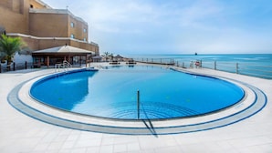 A large, clear swimming pool with a view of the ocean, surrounded by a building with outdoor seating and a palm tree.