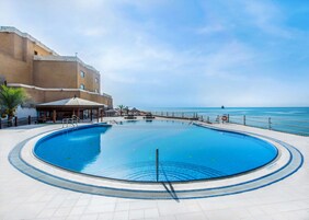 Una piscina amplia y cristalina con vista al océano, rodeada por un edificio con asientos al aire libre y una palmera.