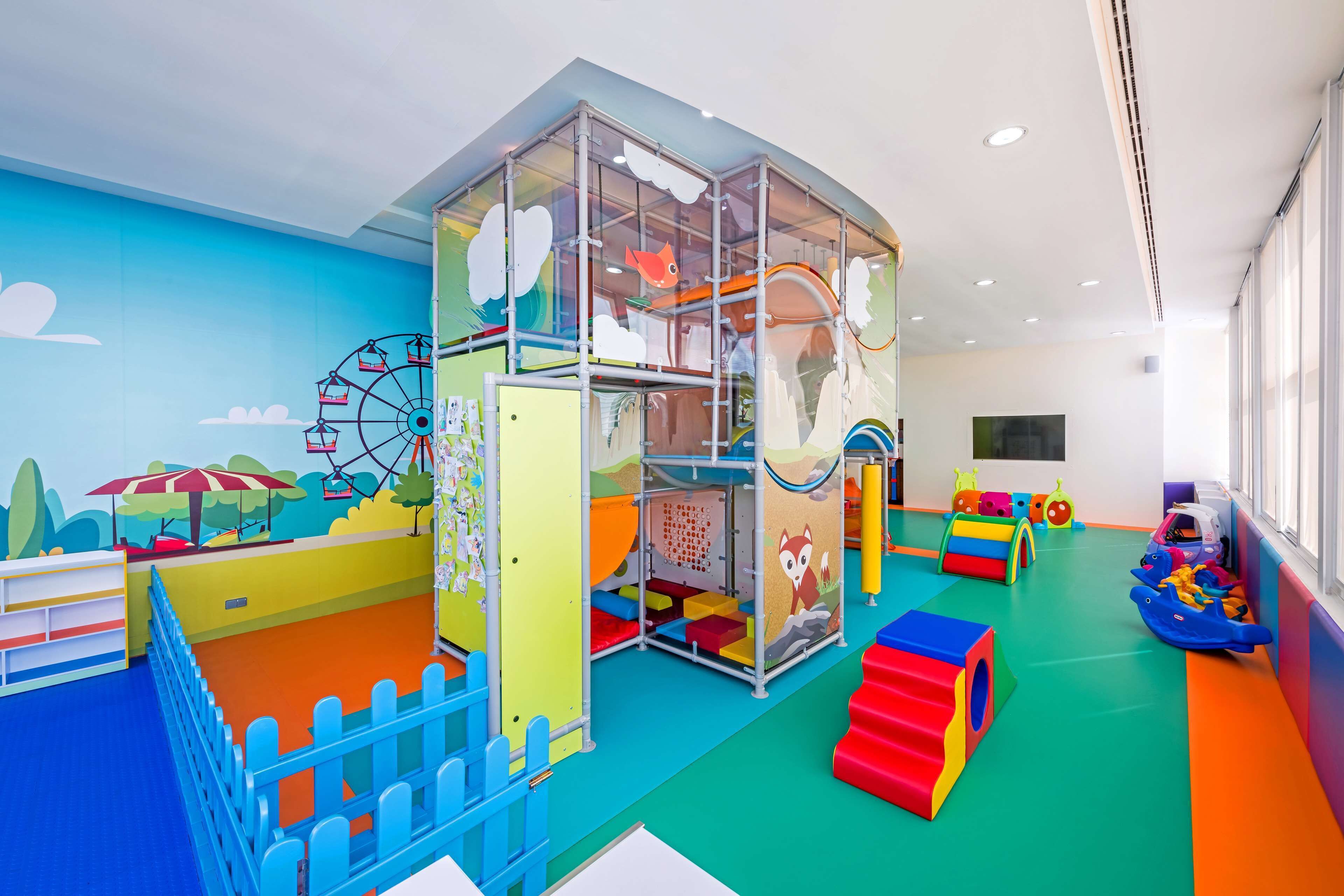 Zona de juegos infantiles con una estructura de escalada, alfombras de colores y un mural de noria.