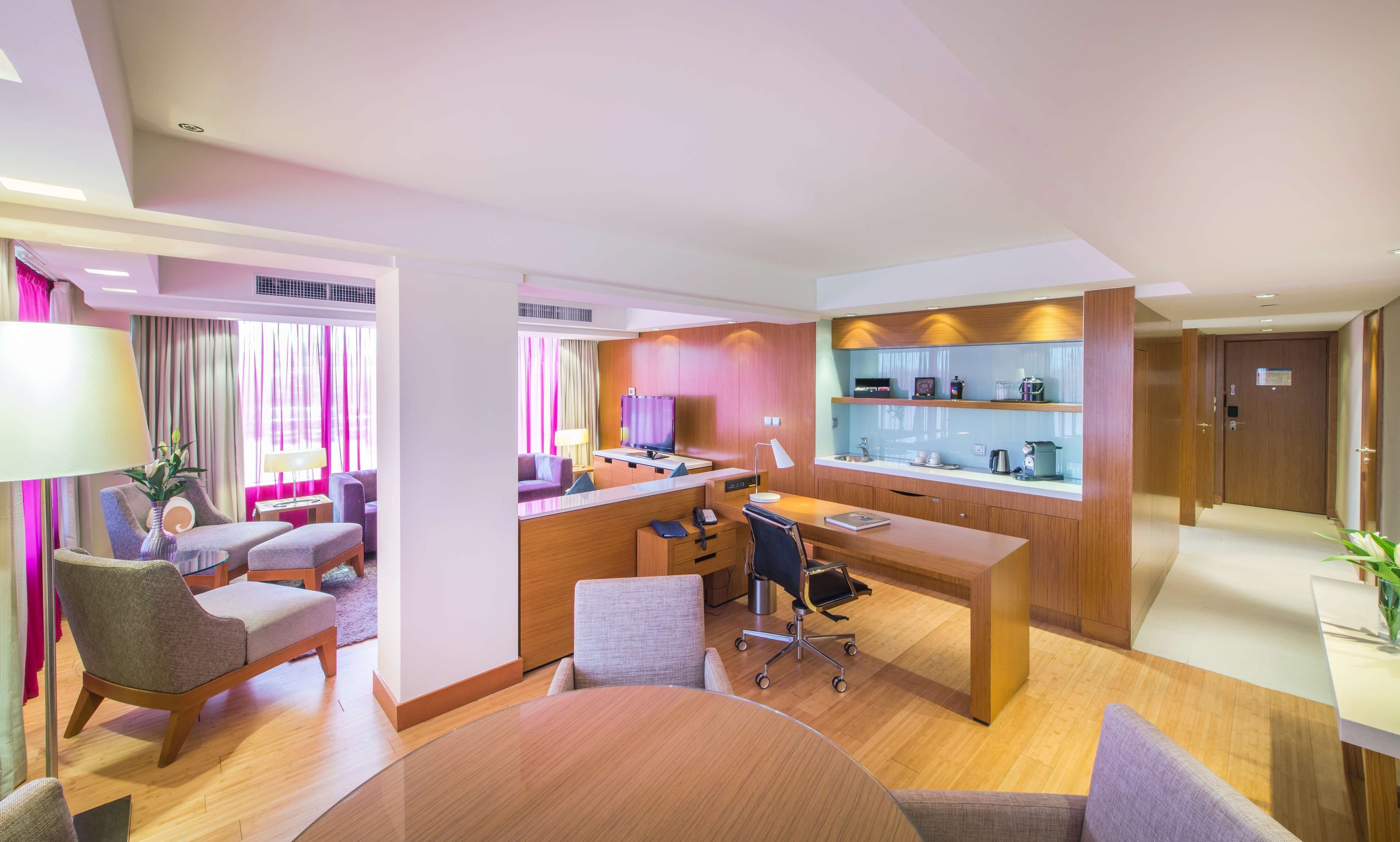 A modern hotel room with a dining area, a sofa set, a desk with a computer, and a coffee machine.