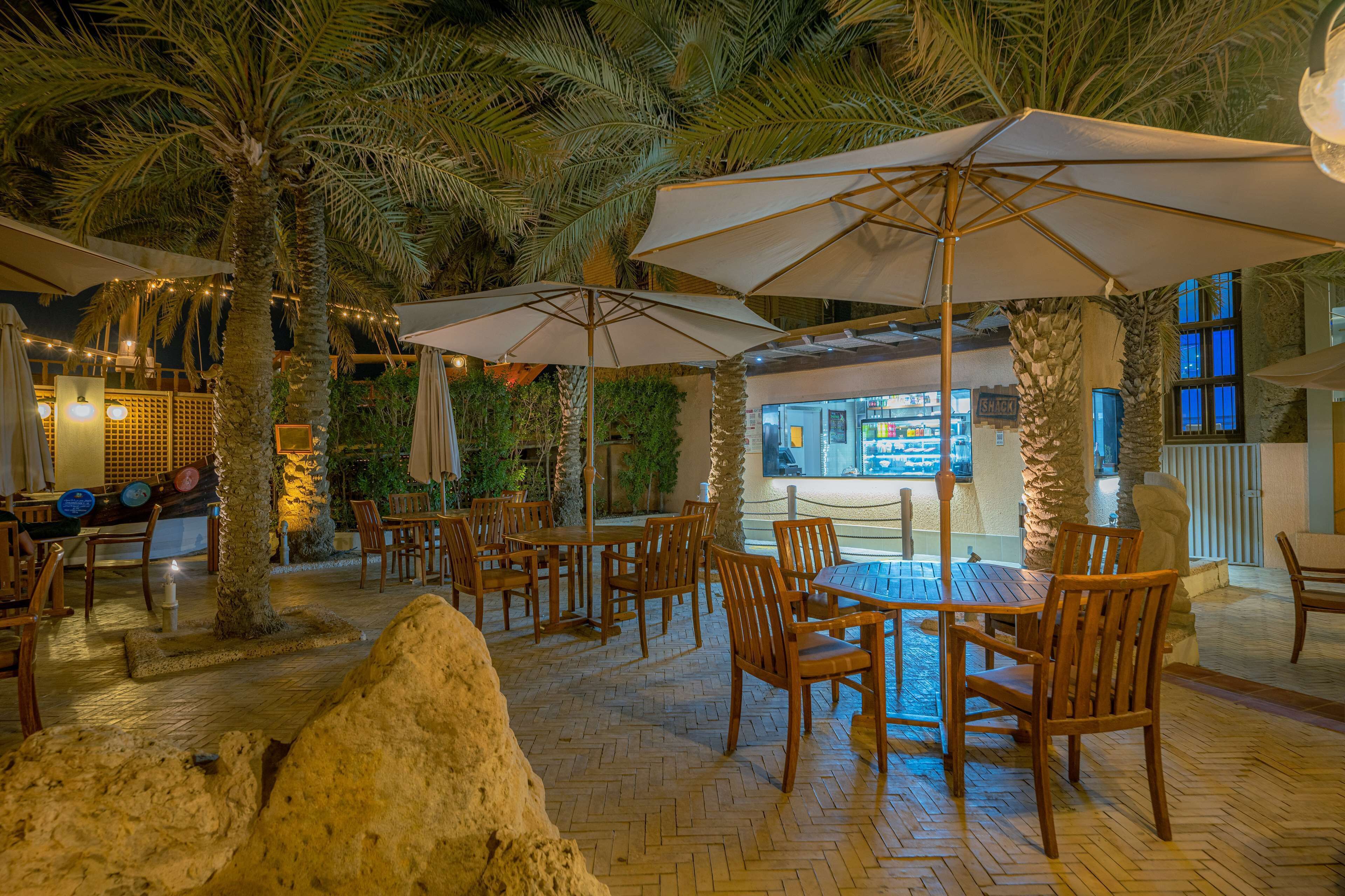 An indoor patio area with wooden tables and chairs, palm trees, and umbrellas.