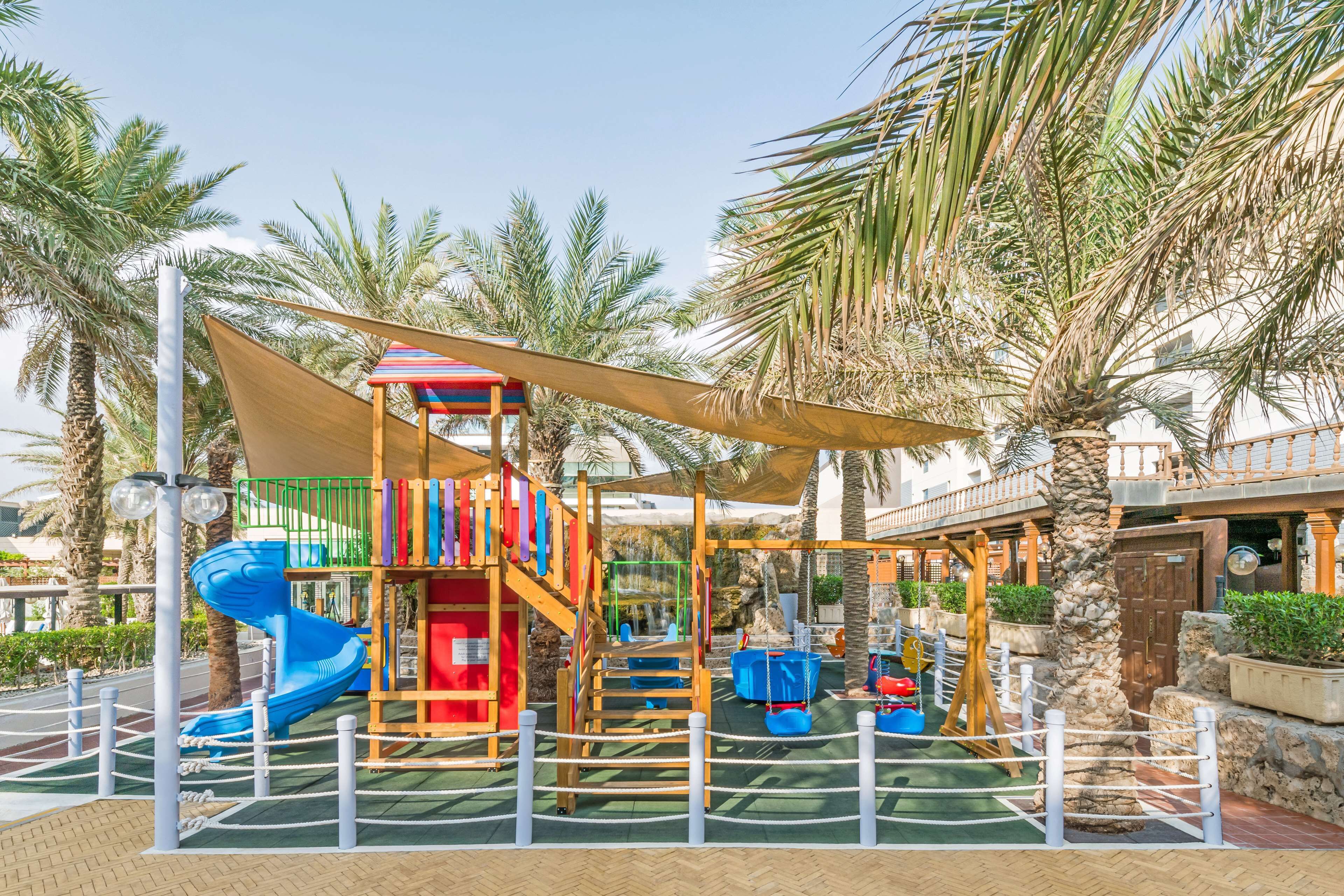 Un parque infantil con tobogán, estructuras para escalar y una zona con sombra.