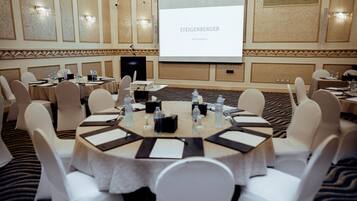 Un salón de conferencias con una pantalla grande que muestra “STEIGENBERGER CECILOTEL”, mesas redondas con sillas, botellas de agua y tarjetas de identificación.