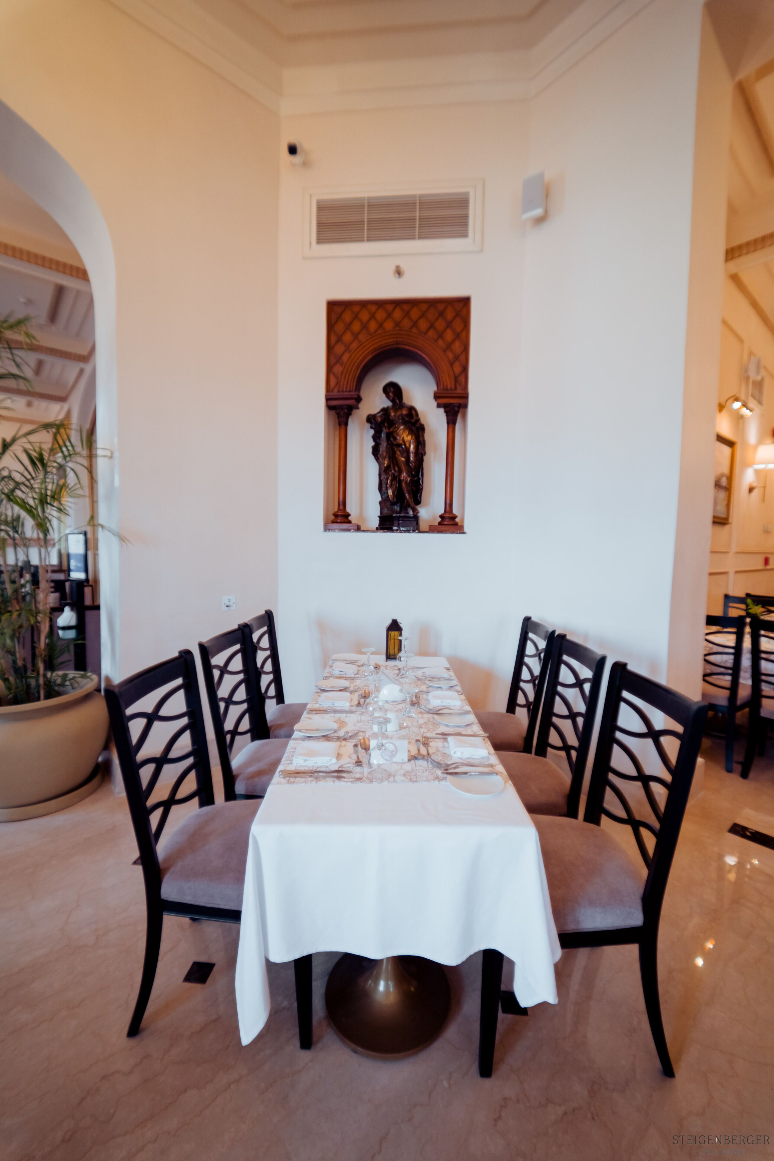 A dining area with a long table set for four, surrounded by chairs, a potted plant, and a decorative wall piece.