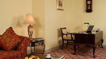 Una sala de estar con un sofá, una mesa baja con frutas, una lámpara, una silla, un escritorio con una computadora portátil y un reloj en la pared.