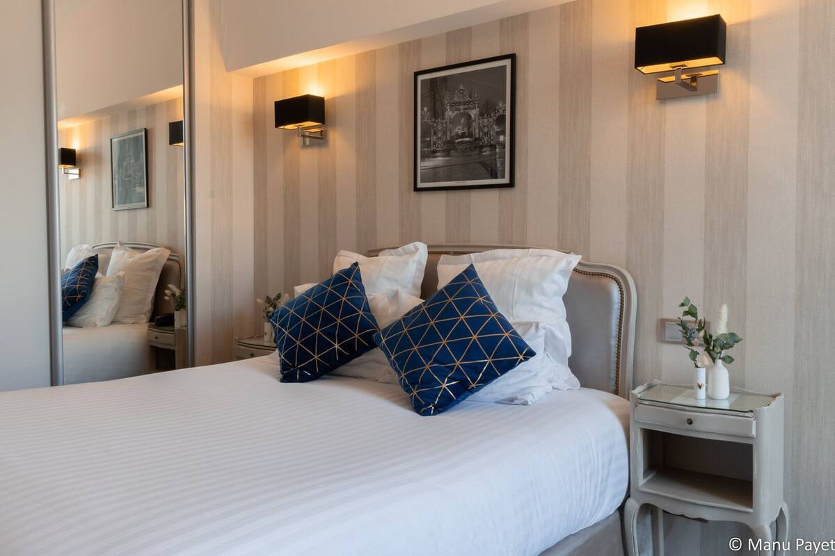 A neatly made bed with white bedding and two blue pillows with gold patterns, a bedside table with a vase of flowers, and a framed picture on the wall.