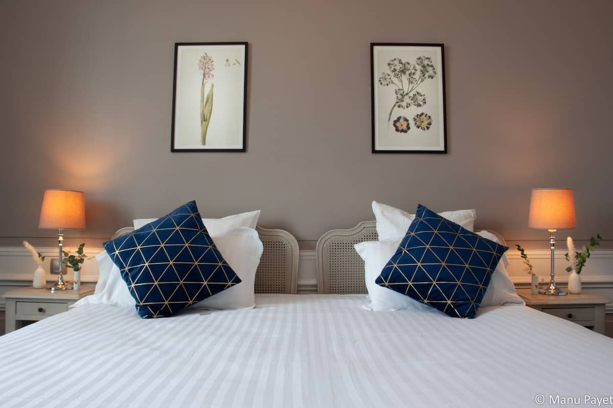 A double bed with white bedding and blue pillows with gold patterns, flanked by two bedside tables with lamps and small vases.