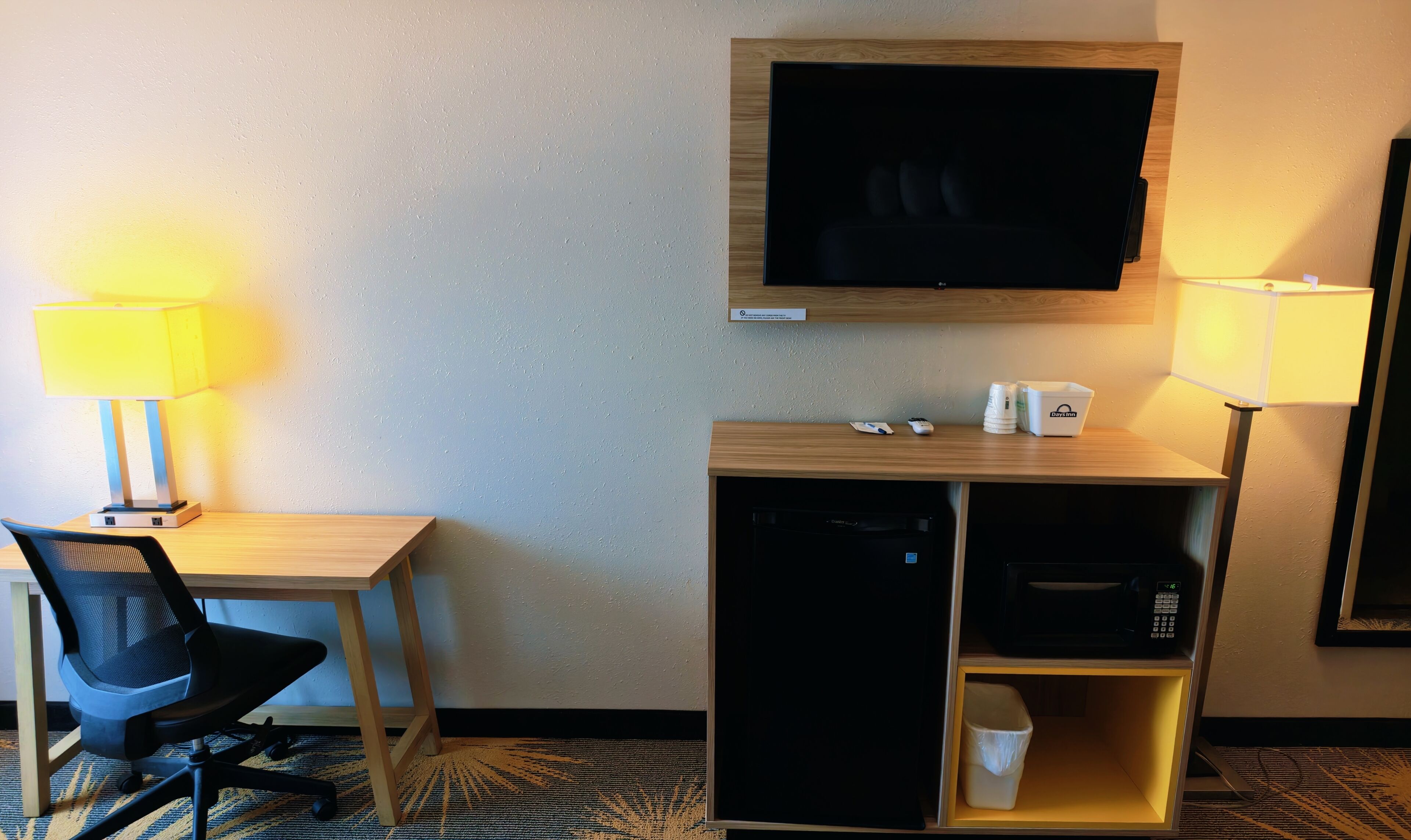 A hotel room with a desk, chair, television, lamps, and a minibar.