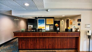 A hotel reception desk with a wooden finish, a digital clock, a telephone, and a sign indicating 'Drop Keys Here'.