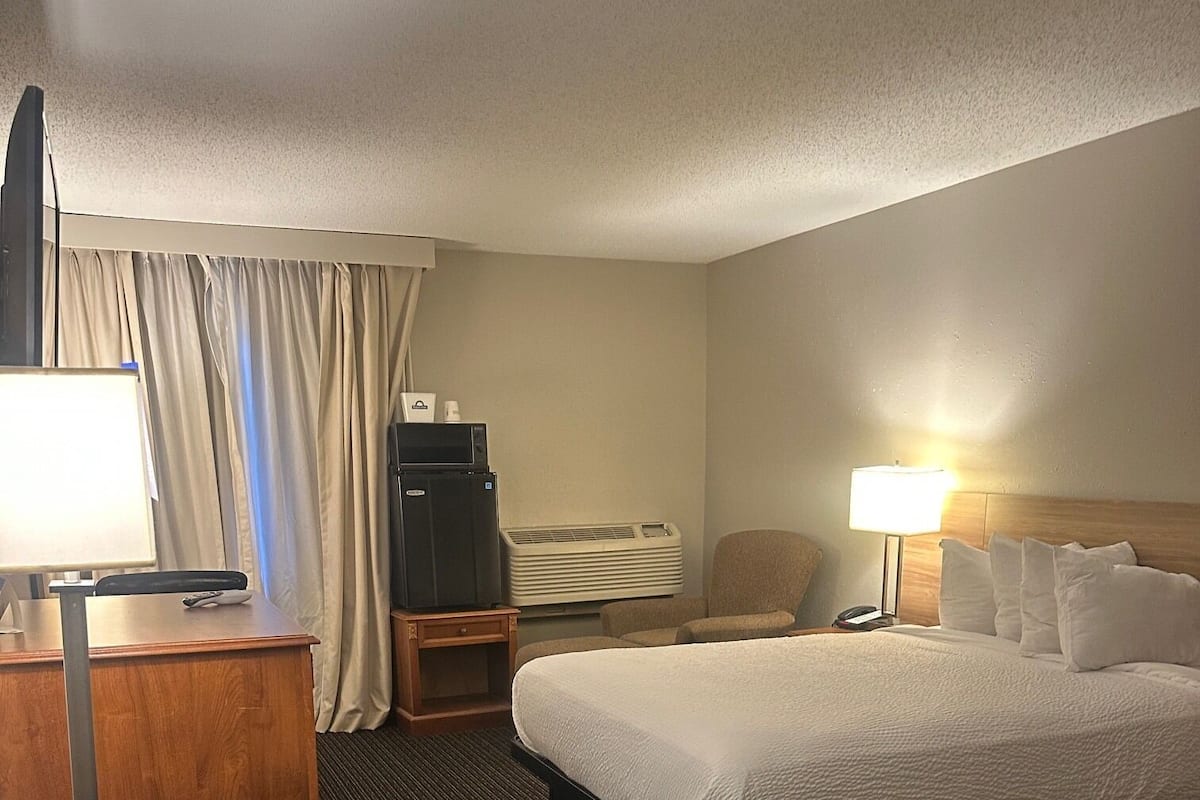 A hotel room with a bed, a desk with a lamp, a chair, an air conditioning unit, and a television.