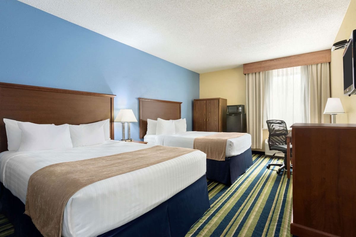 A hotel room with two beds, a desk, a chair, a microwave, and a television.