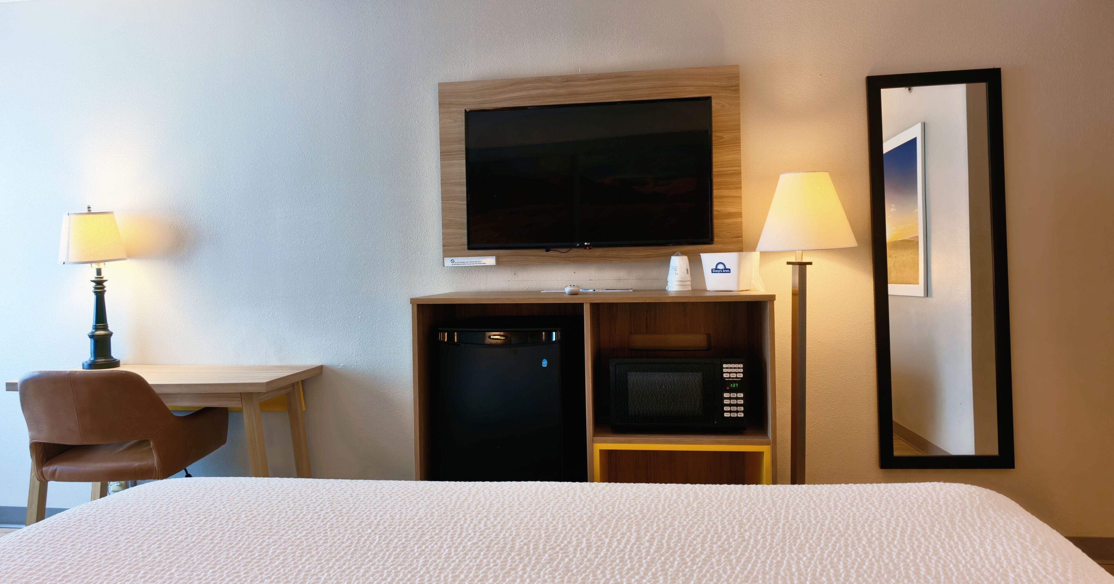 A hotel room with a bed, a television, a microwave, a desk with a chair, and two lamps.