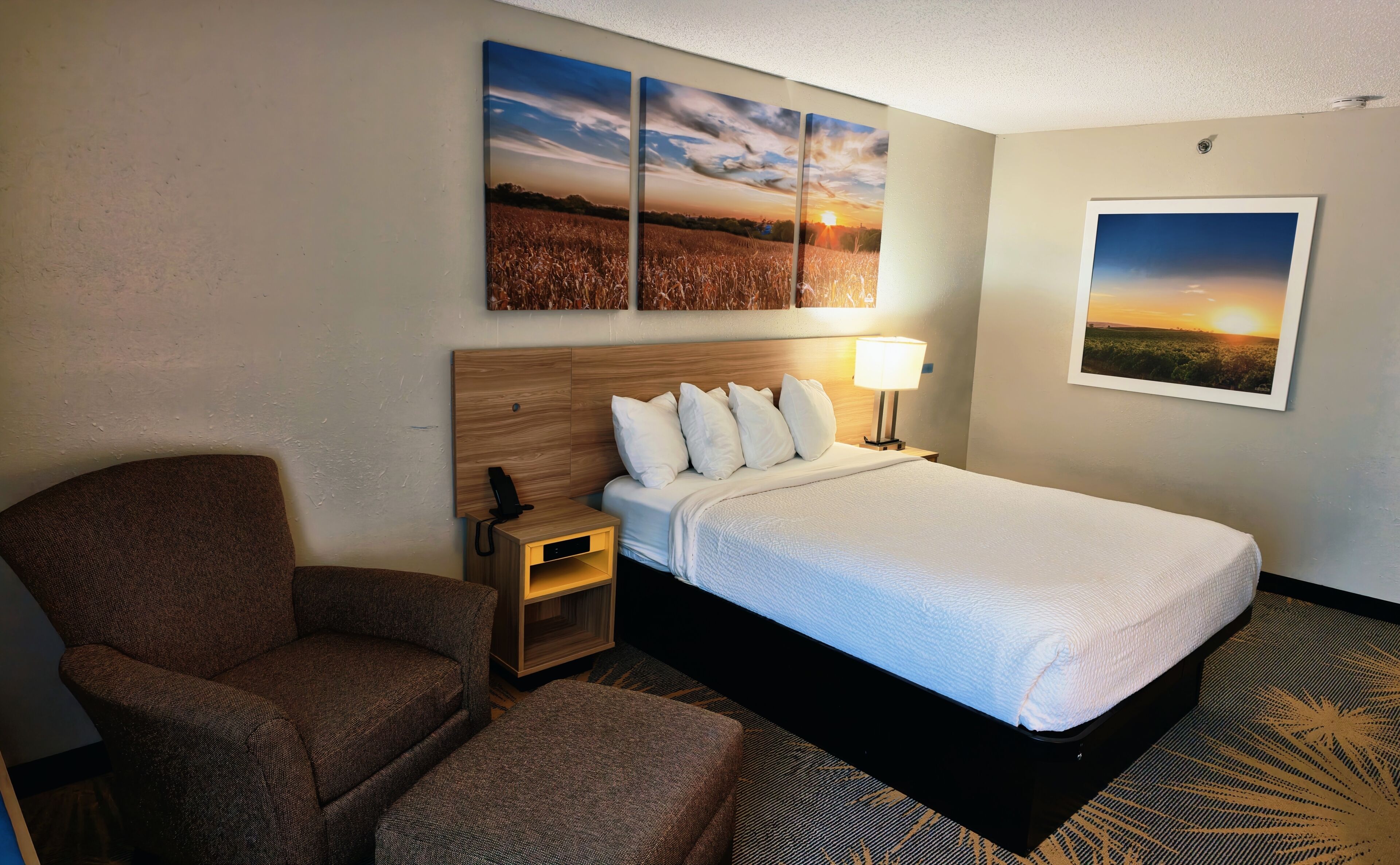 A hotel room with a bed, a chair, a nightstand, a lamp, and wall art depicting landscapes.