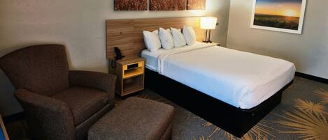 A hotel room with a bed, a chair, a nightstand, a lamp, and wall art depicting landscapes.