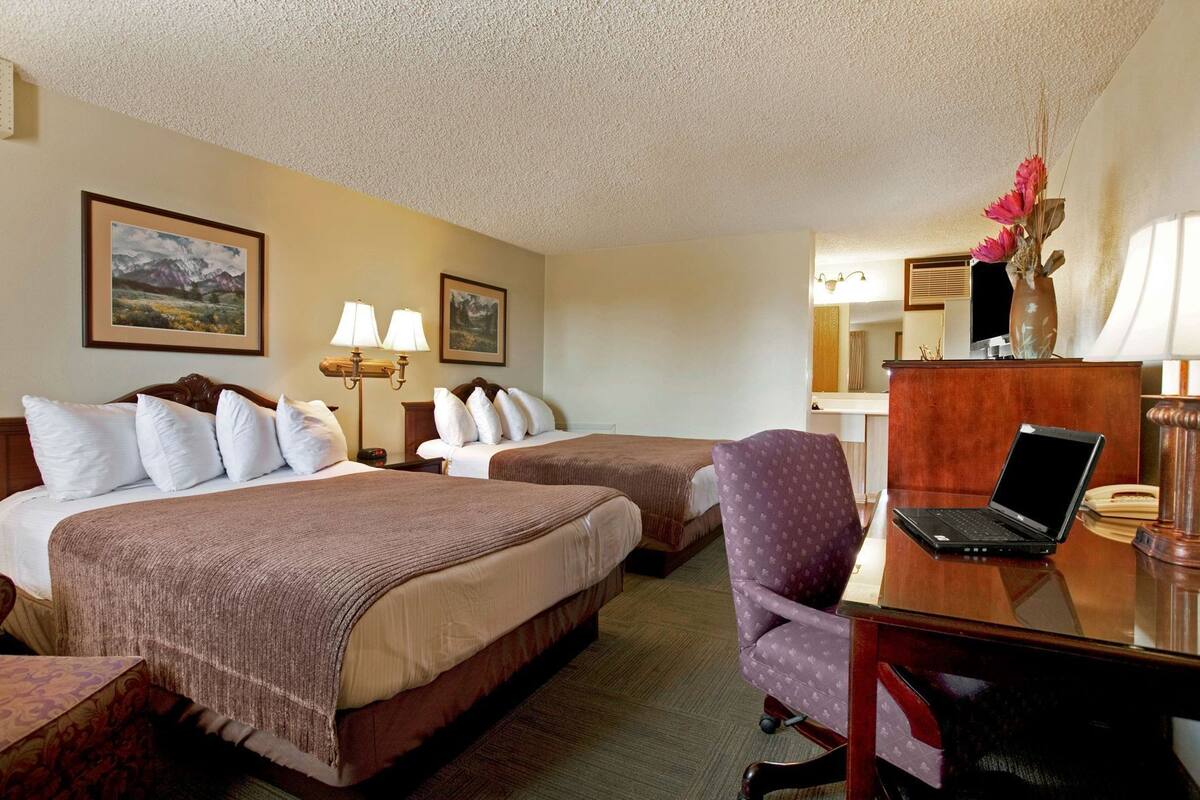 A hotel room with two beds, a desk with a laptop, a chair, a desk lamp, and two wall-mounted lamps.