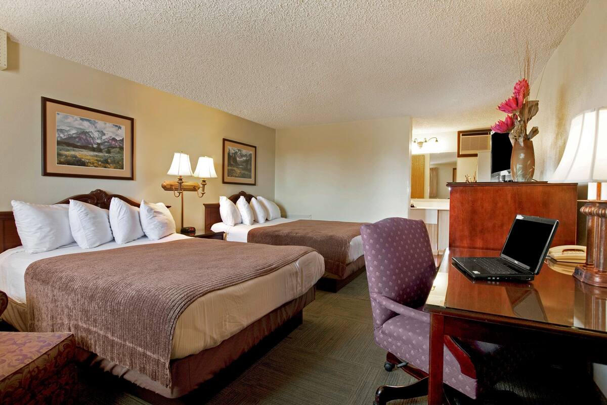 A hotel room with two beds, a desk with a laptop, a chair, and a vase with flowers.