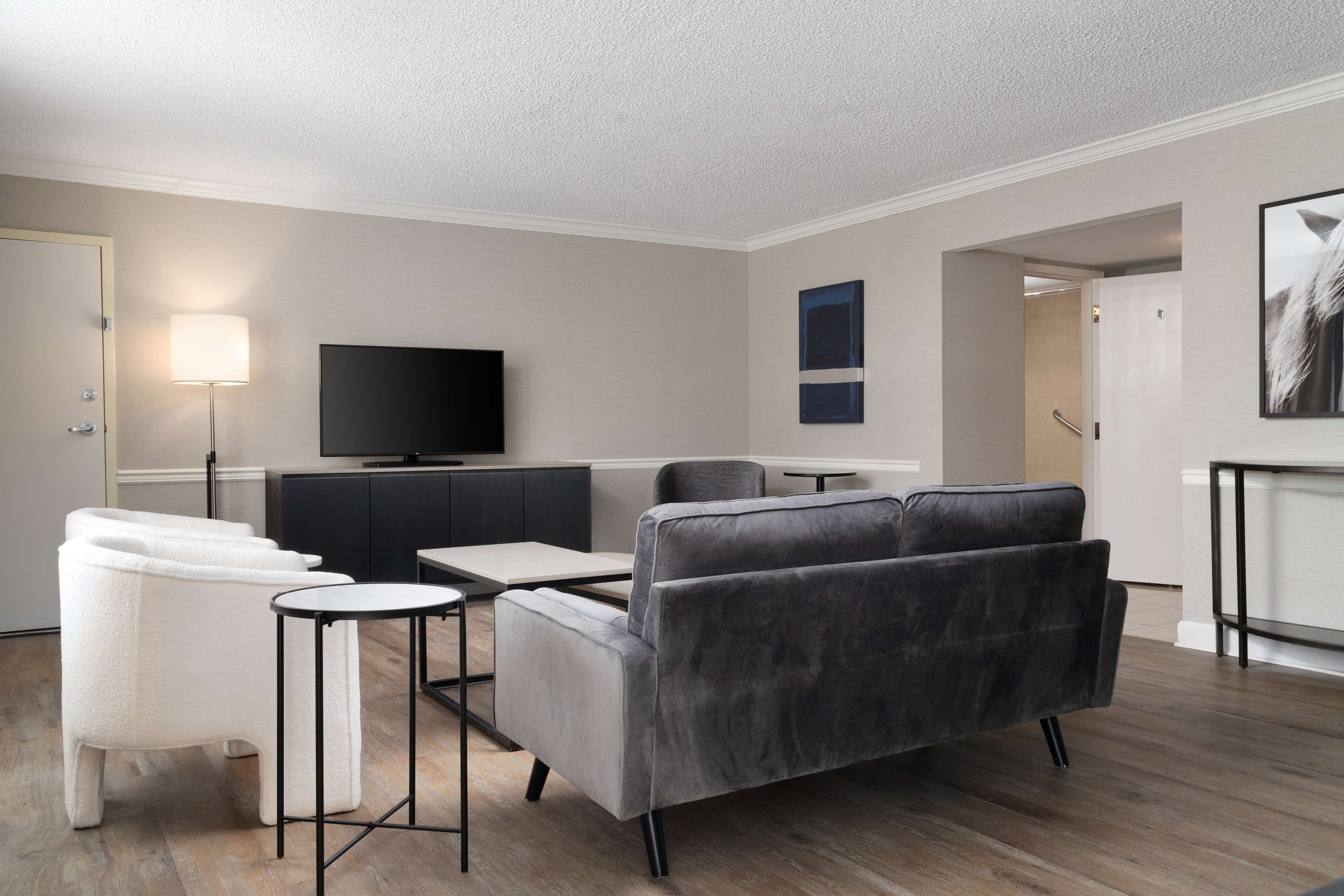 Una sala de estar moderna con un sofá gris, un sillón blanco y un televisor de pantalla plana. Hay una mesita y una lámpara. El suelo es de madera y hay una puerta que da a otra zona.