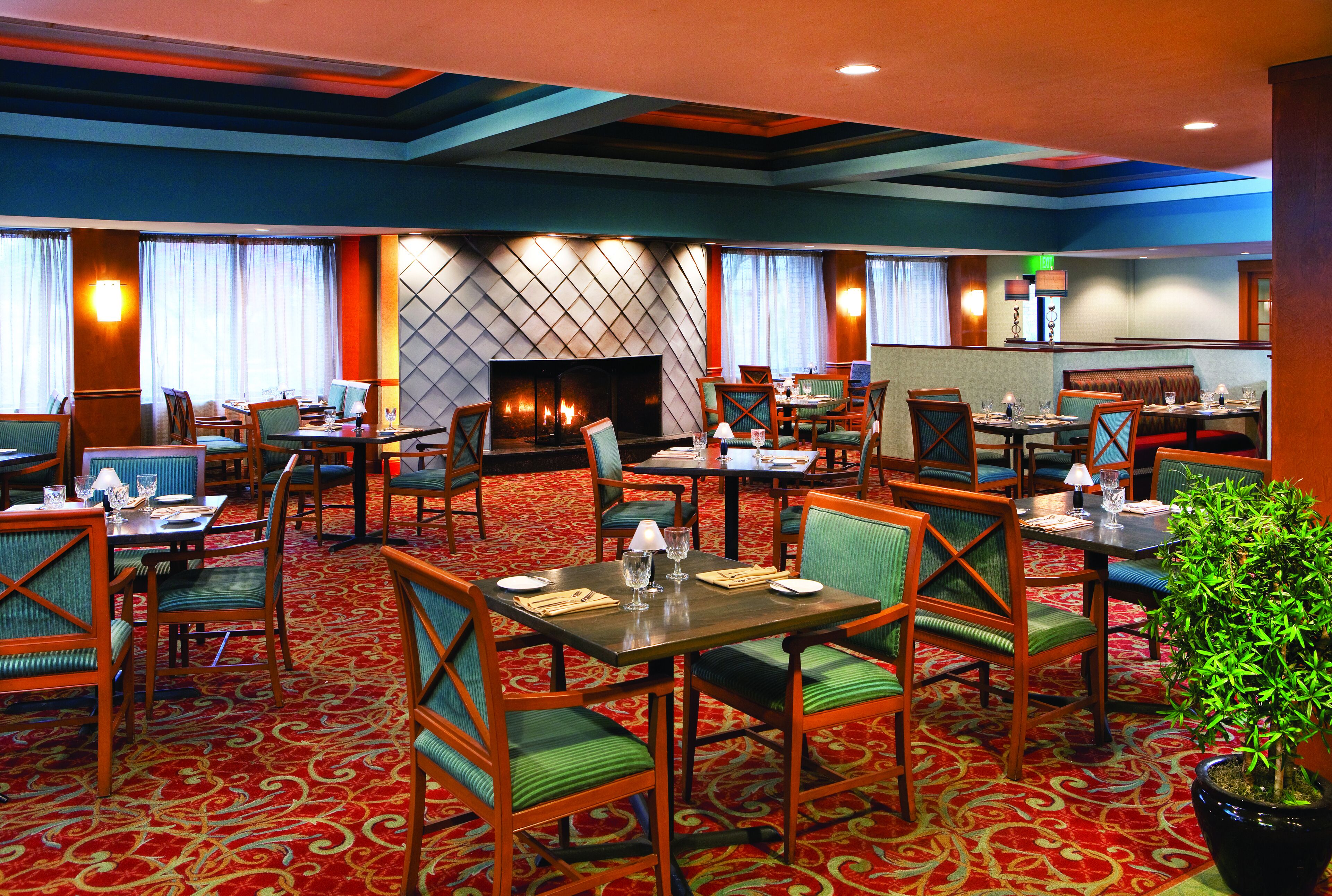 A hotel dining area with multiple tables and chairs, a fireplace, and a patterned carpet.