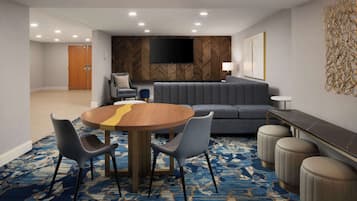 Une chambre d’hôtel moderne avec une table de salle à manger ronde, un canapé gris recouvert de tissu et un téléviseur à écran plat fixé à un mur en bois.