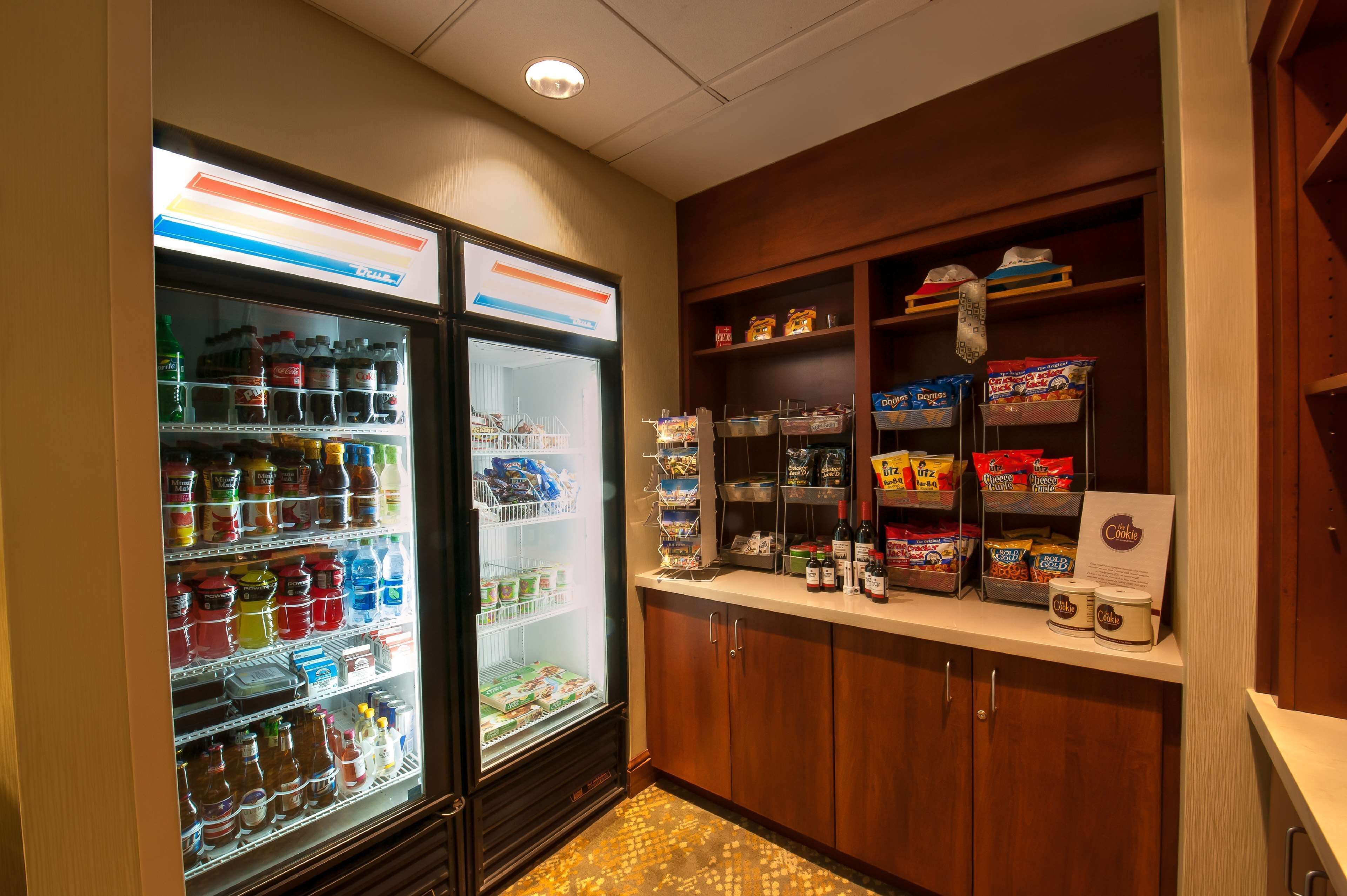 A hotel pantry with two refrigerators stocked with beverages and snacks, and shelves with packaged goods and condiments.