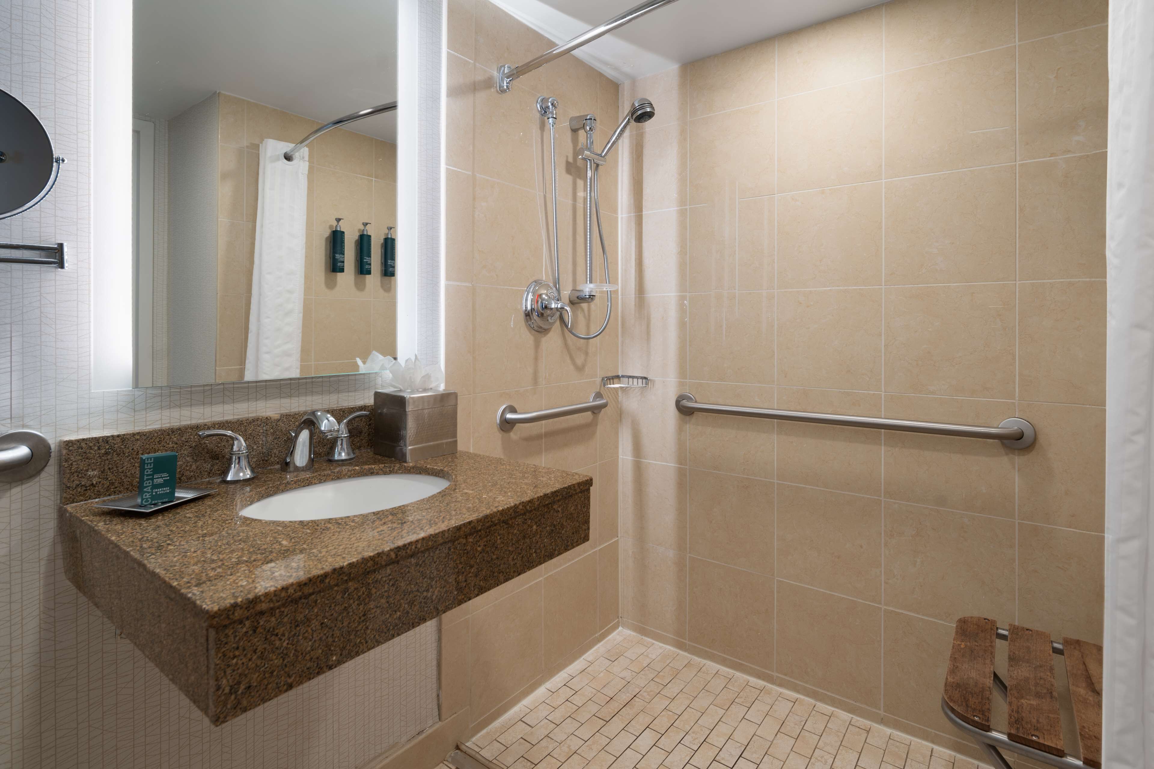 A bathroom with a granite countertop sink, a mirror, a shower with a handheld showerhead, and a grab bar.
