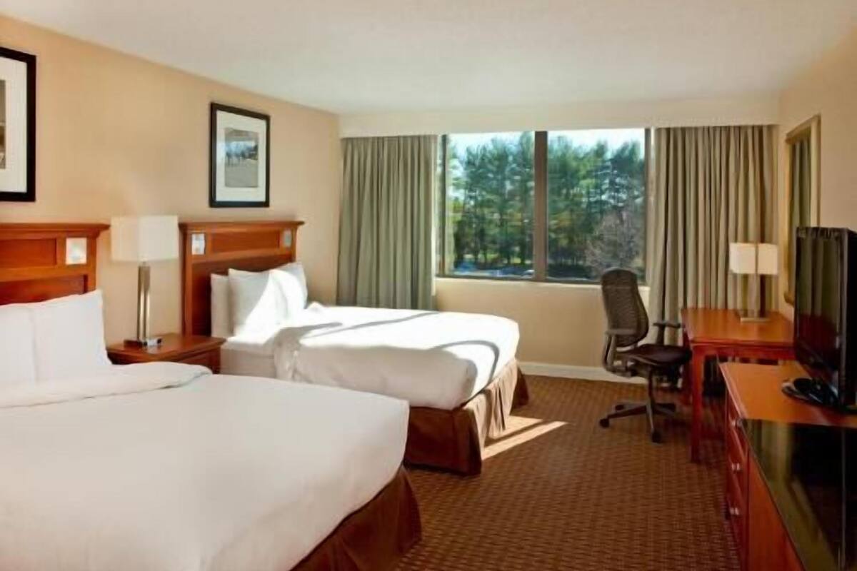 A hotel room with two beds, a desk with a television, a chair, and a window with curtains.