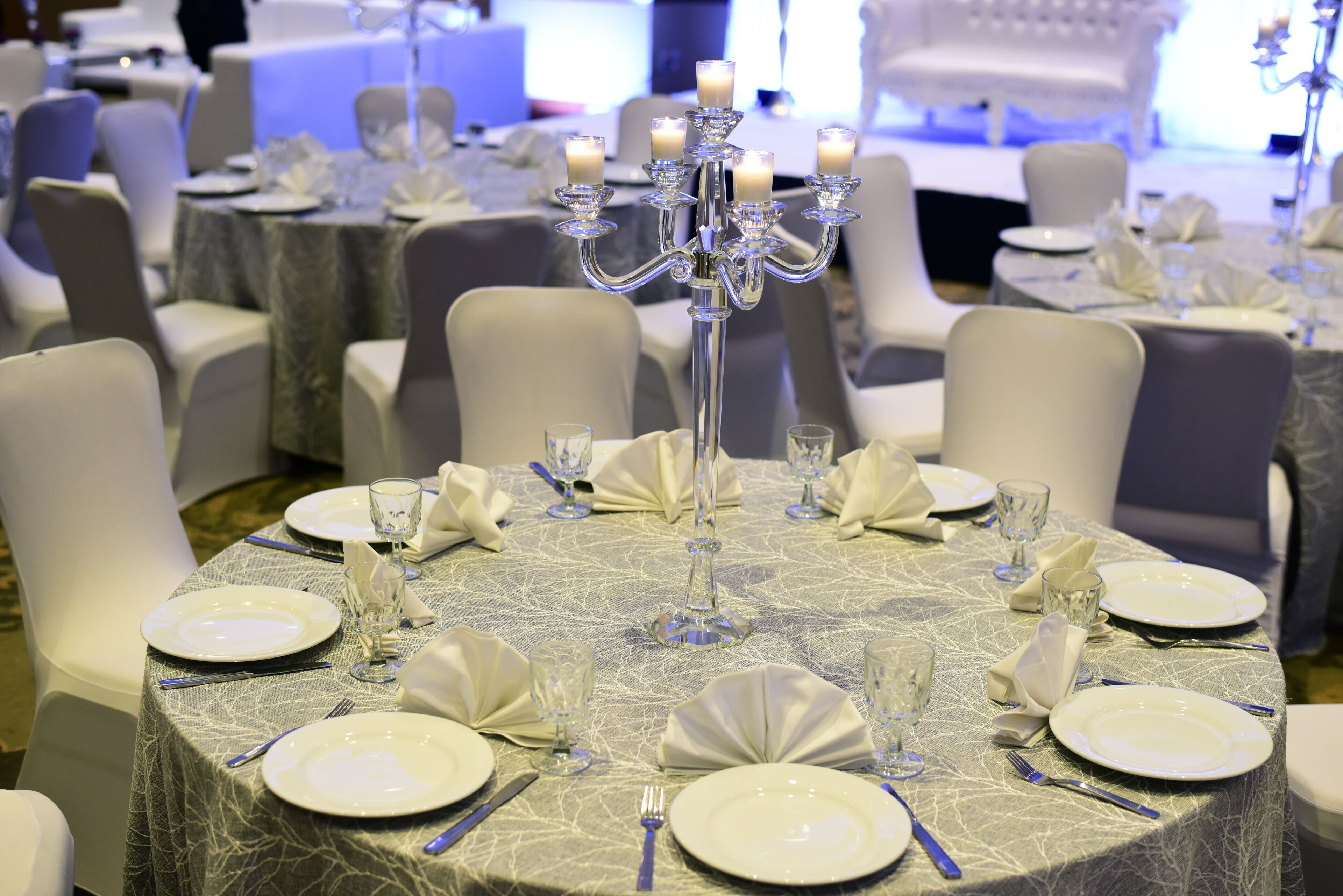Une table ronde dressée pour un événement formel, avec de l’argenterie, de la vaisselle blanche et des verres, entourée de chaises blanches.