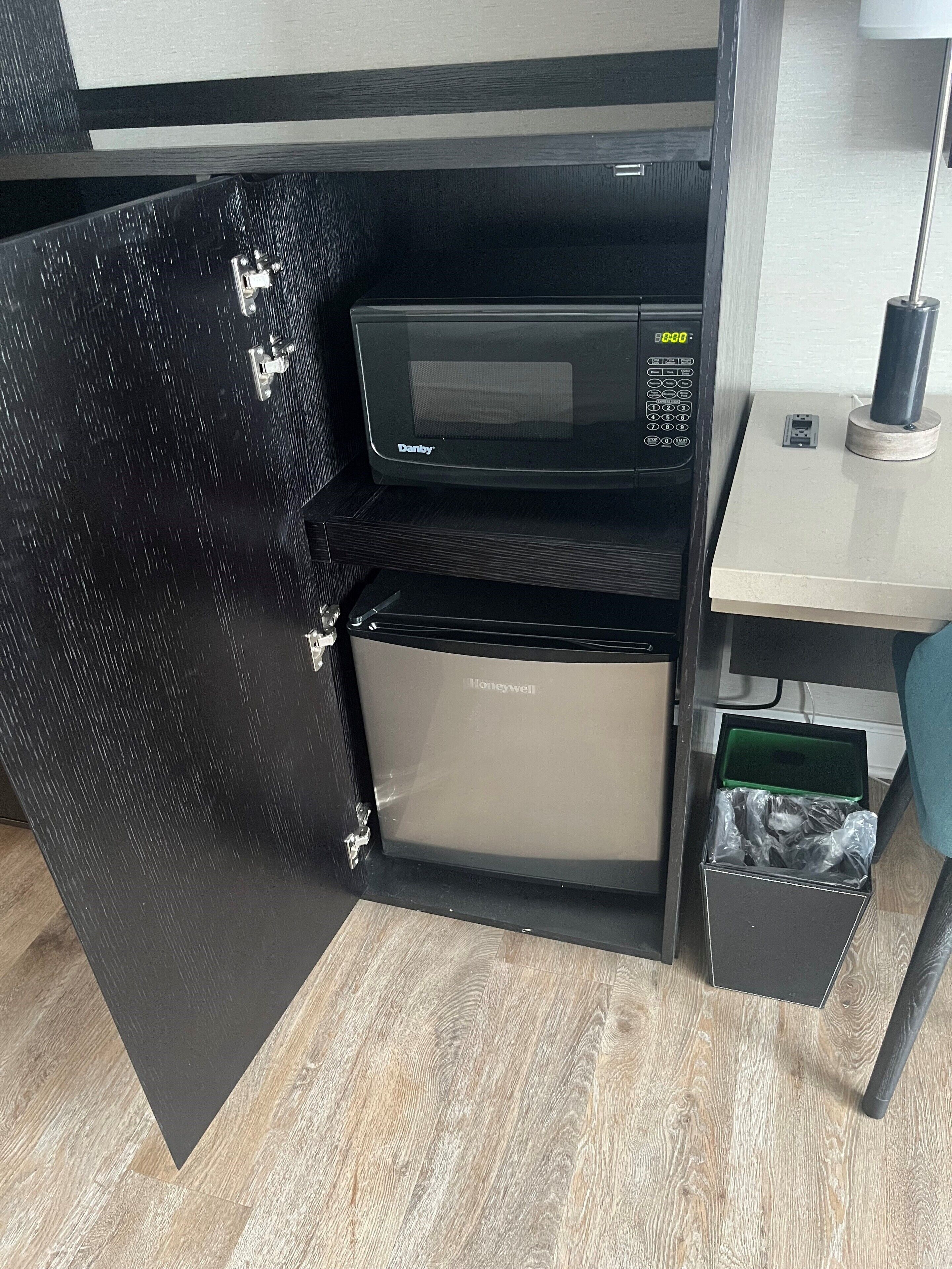 A compact kitchen with a microwave and a refrigerator, a small trash bin, and a power outlet on the countertop.