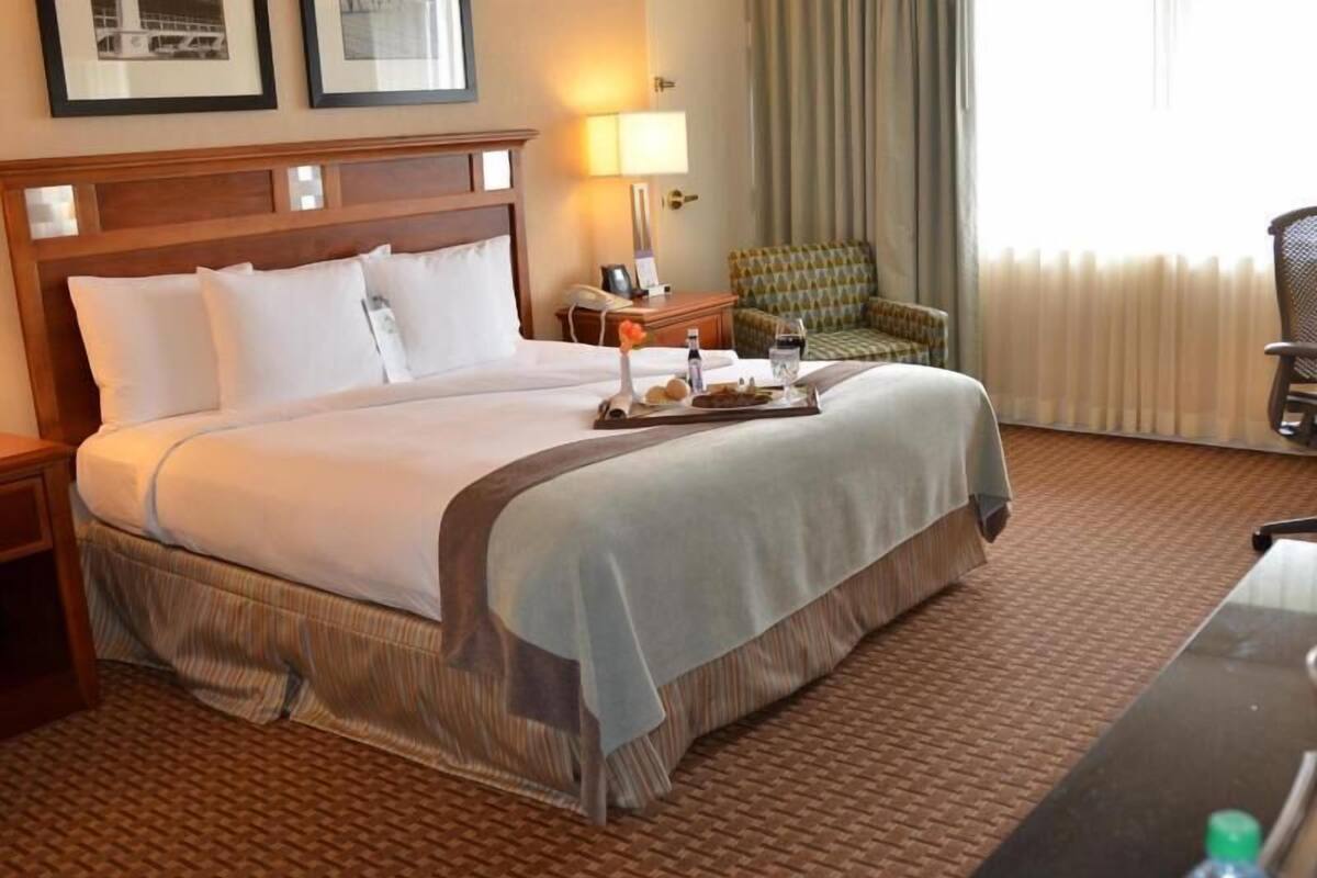 A hotel room with a large bed, a bedside table with a lamp, a chair, and a desk with a bottle.