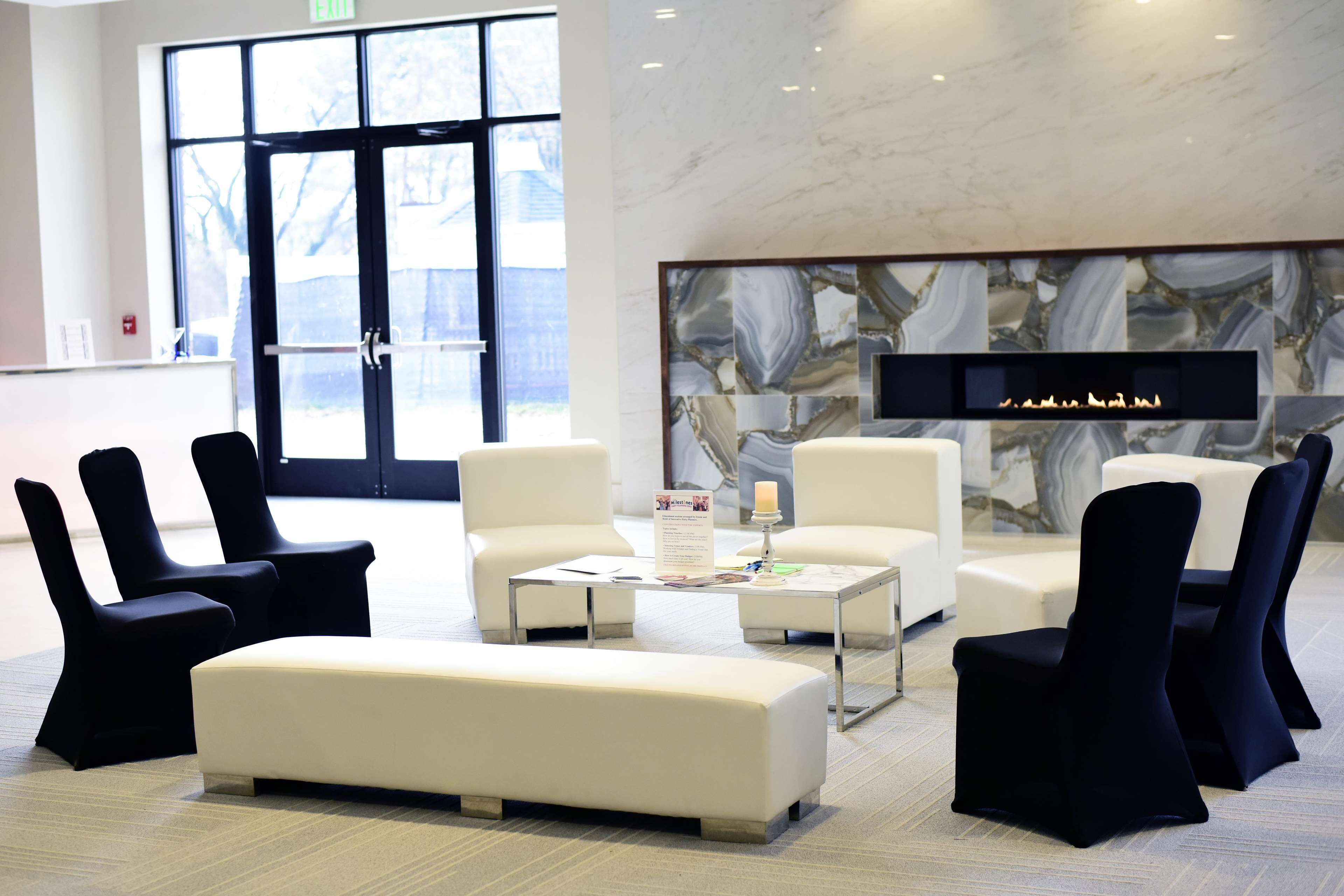 A modern lobby with a marble wall, seating area with white and black chairs, and a large window.