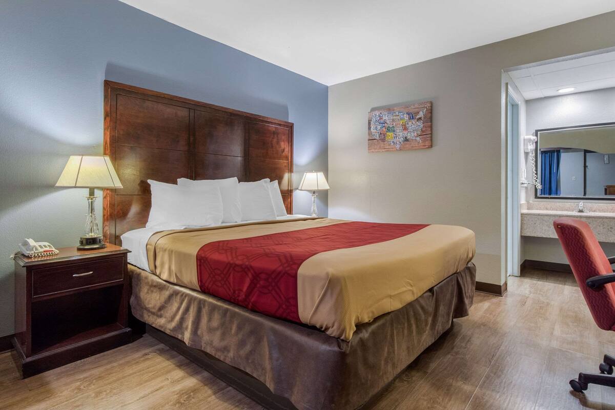 A hotel room with a large bed, two bedside tables with lamps, a wooden headboard, a red and beige bedspread, a small desk with a telephone, and a red office chair.