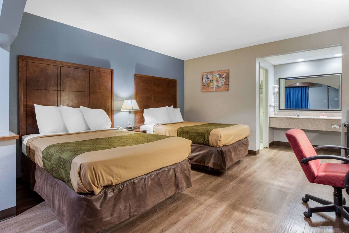 A hotel room with two beds, a desk, a chair, and a bathroom visible through an open door.