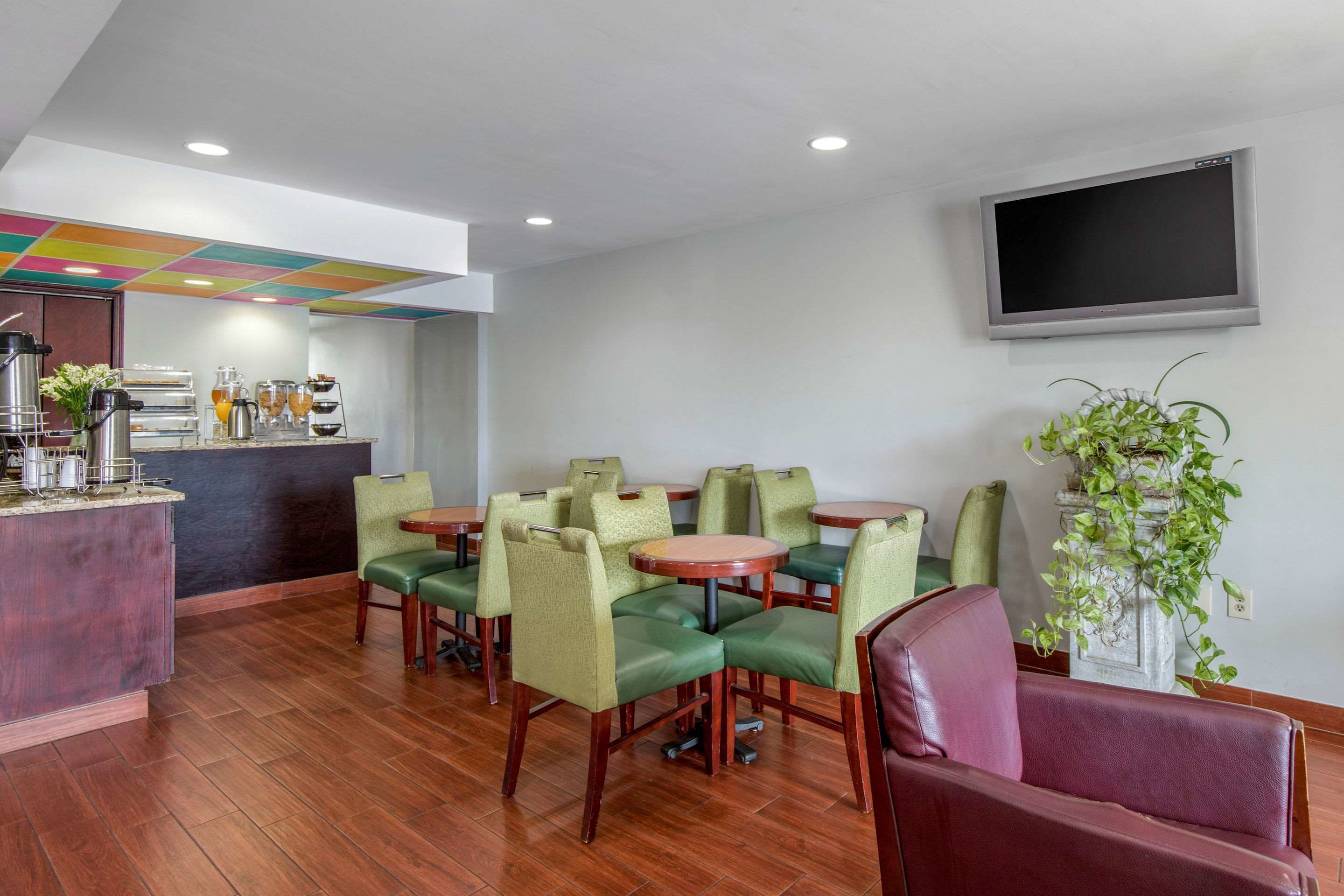 Un hall de hotel con una zona de bar, asientos con sillas tapizadas en verde, un televisor en la pared y una planta en maceta.