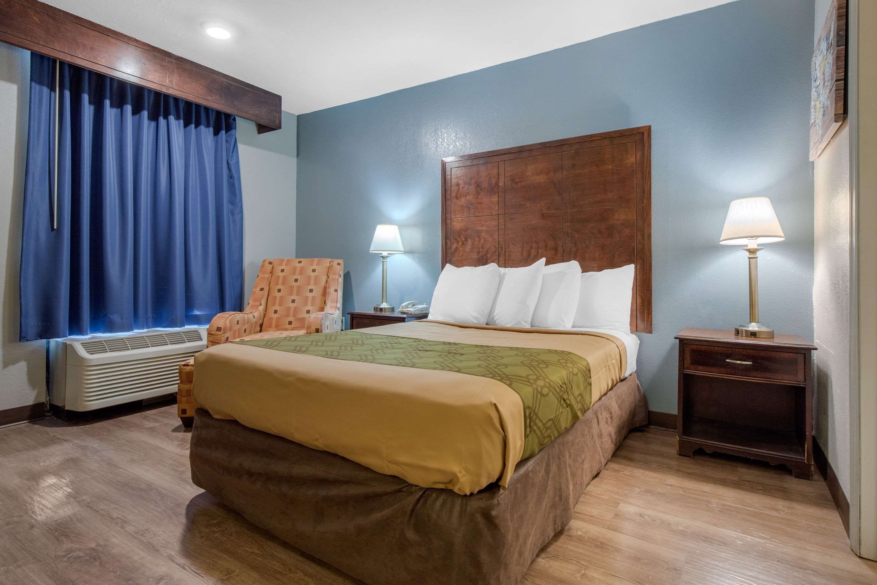 Quarto de hotel com uma cama grande, duas mesas de cabeceira com luminárias, uma poltrona, ar condicionado e uma janela com cortinas.