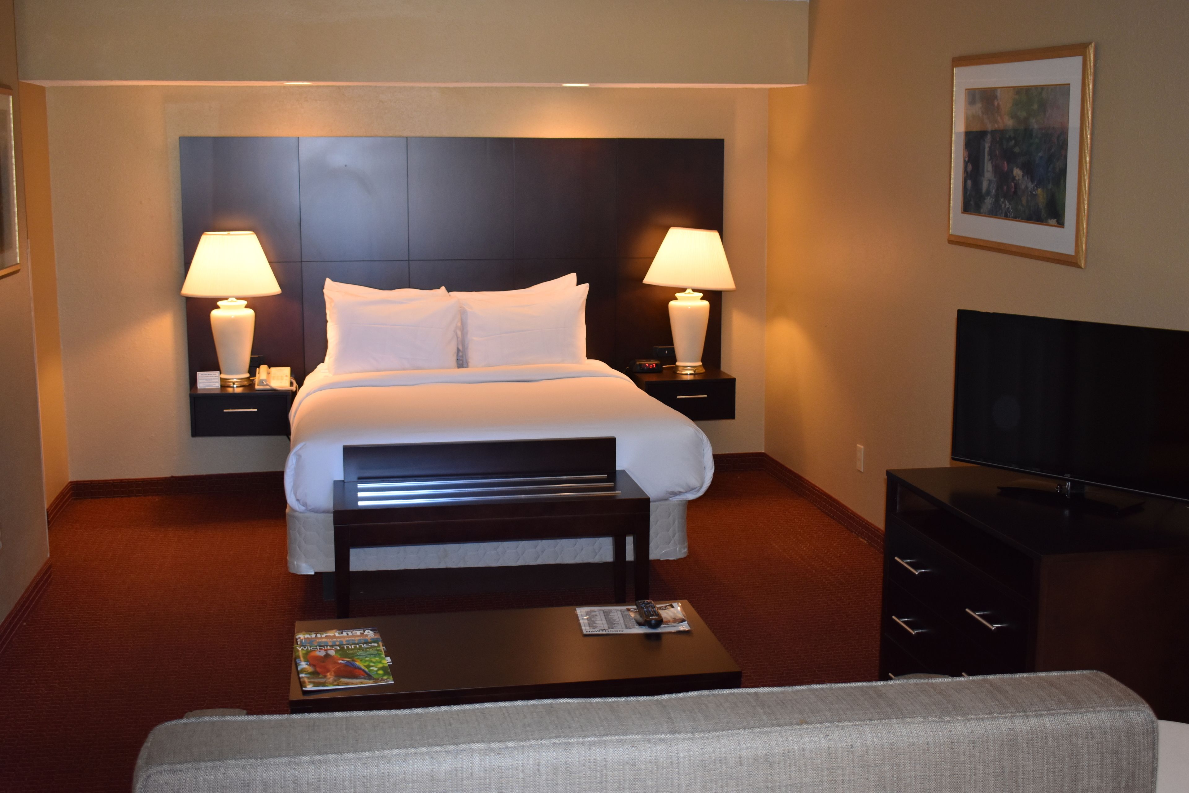 A hotel room with a bed, two bedside lamps, a television, a coffee table, and a framed picture on the wall.