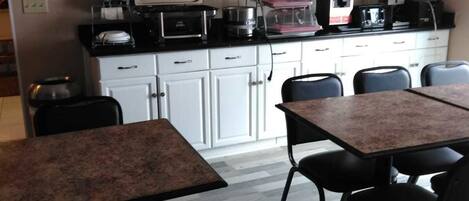 A small dining area with a table and chairs, a kitchen area with appliances, and a framed painting on the wall.