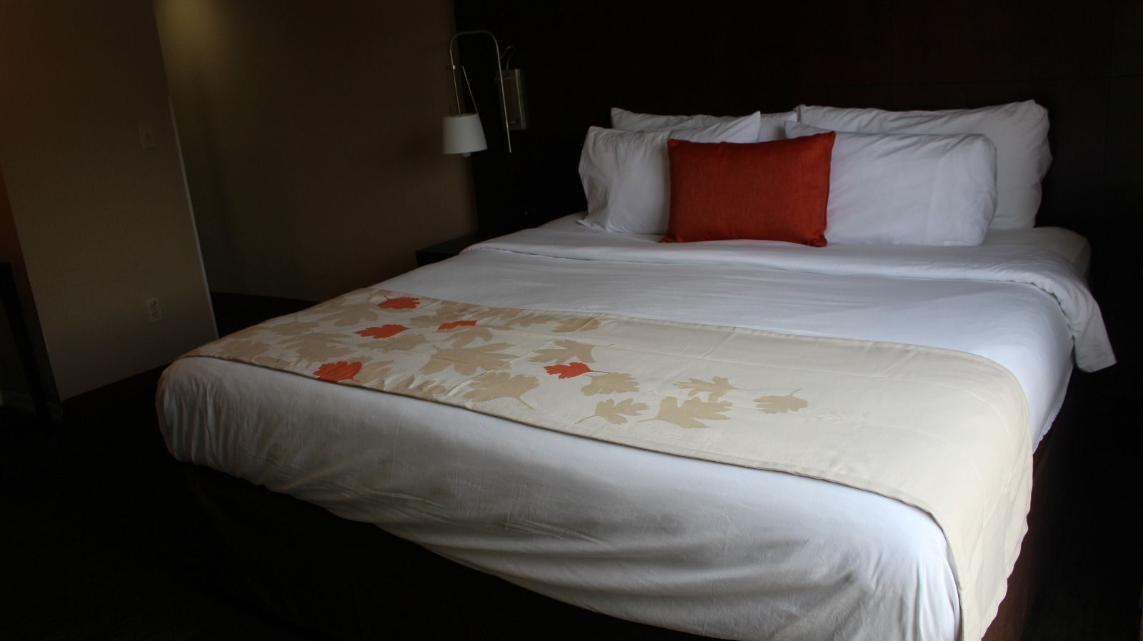 A hotel room with a bed, white bedding, a red pillow, and a wall-mounted lamp.