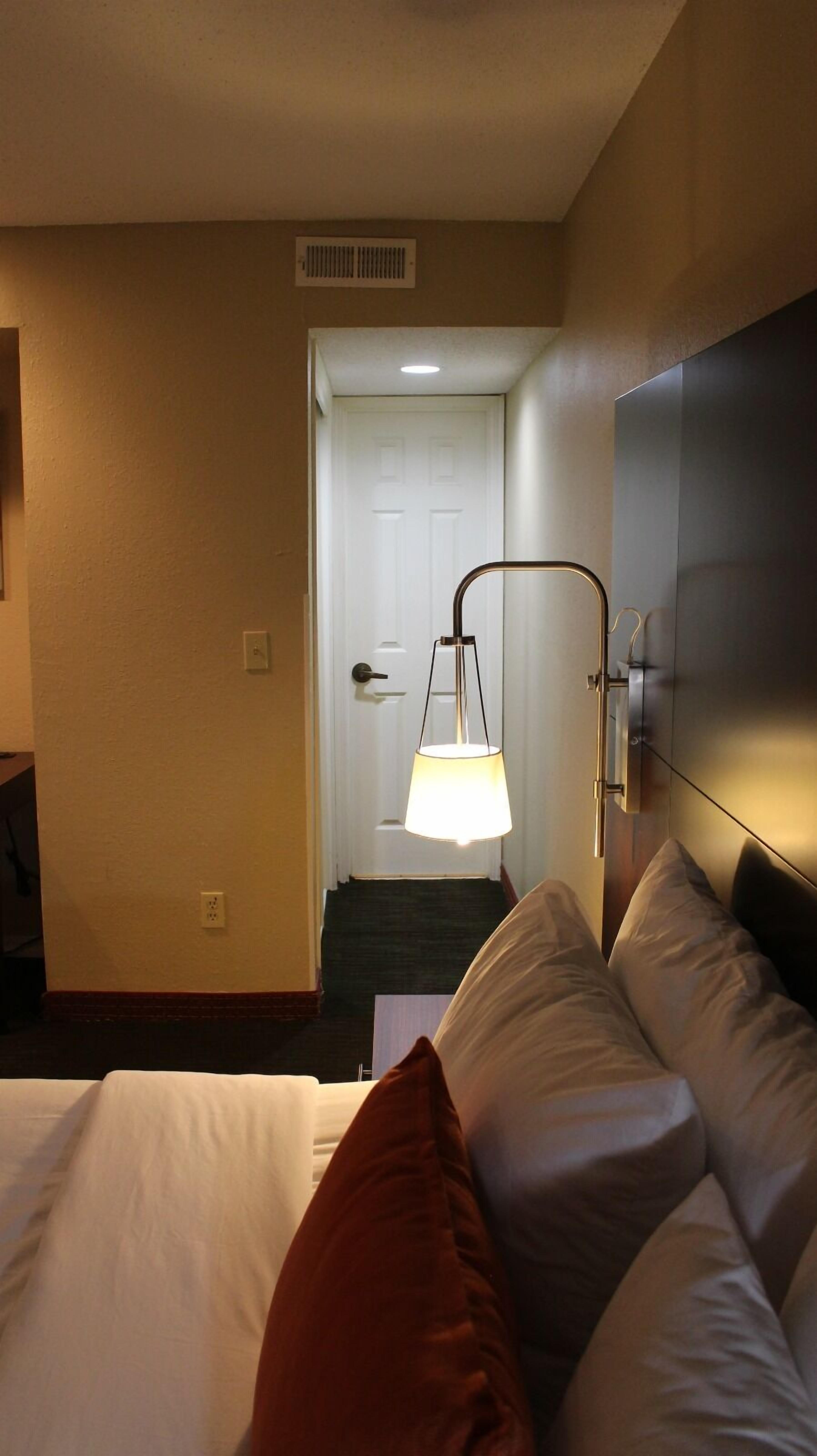 A hotel room with a bed, a bedside lamp, a door, and a wall-mounted panel.