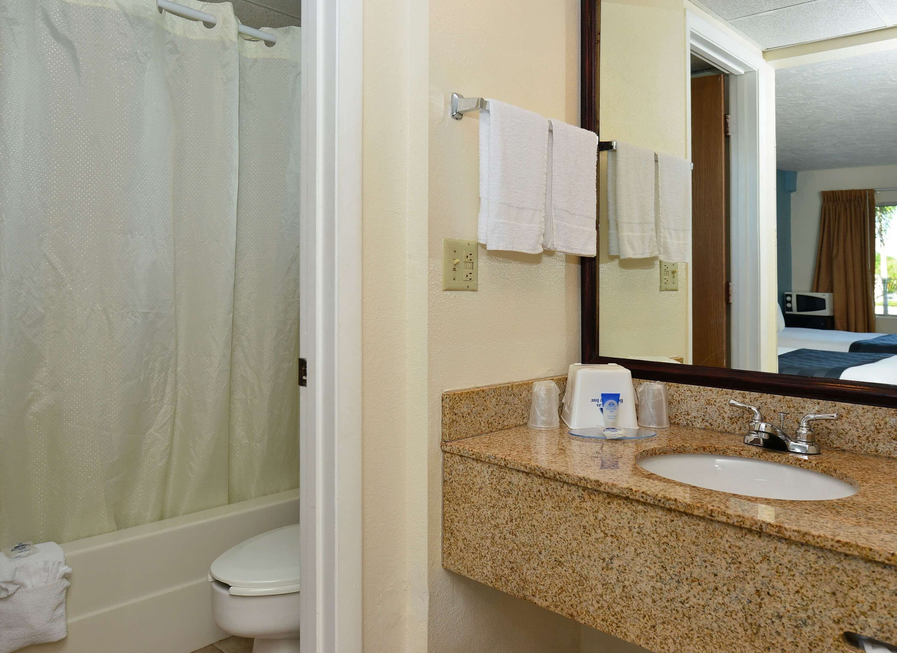 A bathroom with a shower, toilet, sink, and a mirror reflecting a bedroom.