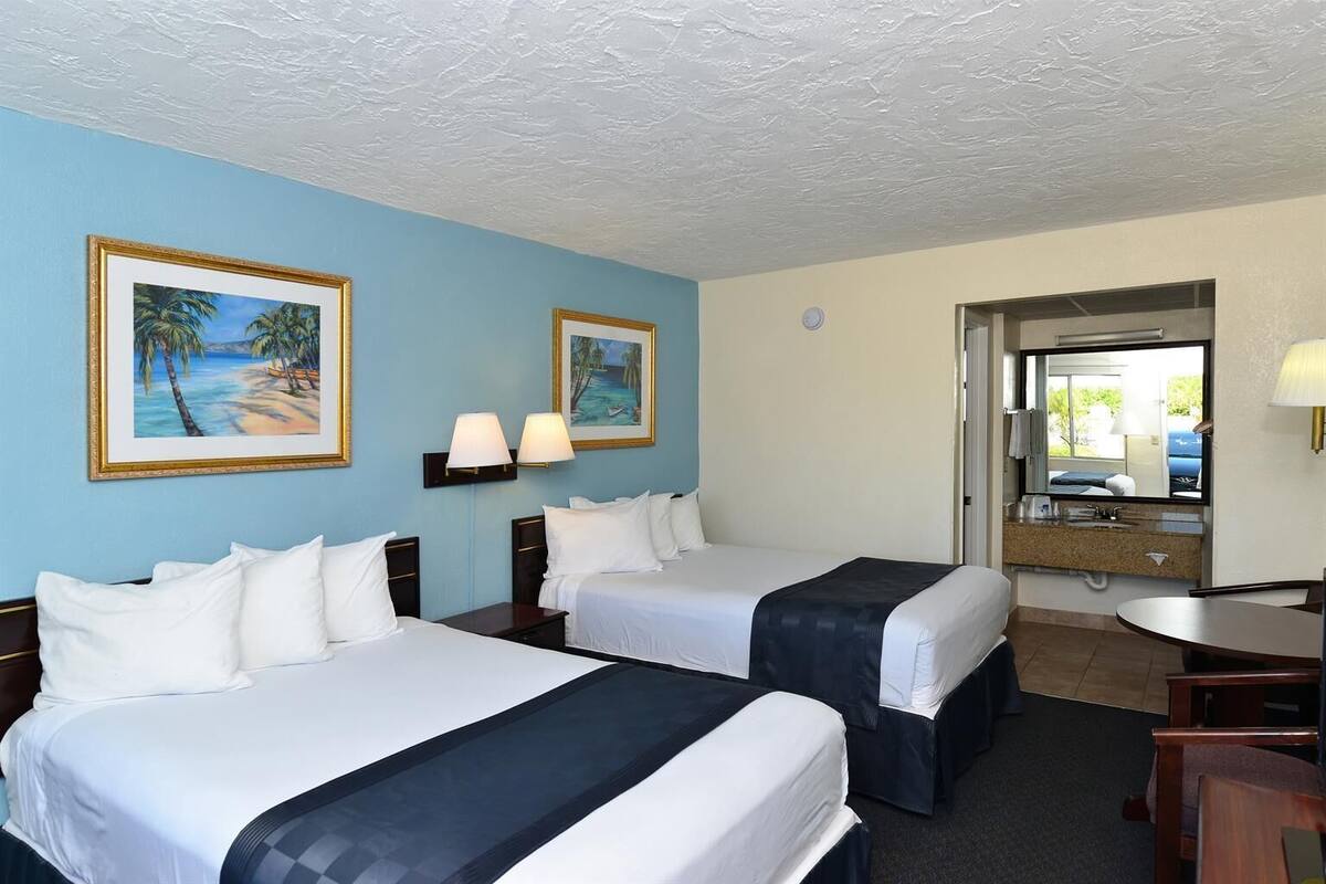 A hotel room with two beds, a desk, a chair, a bathroom mirror, and two wall-mounted lamps.